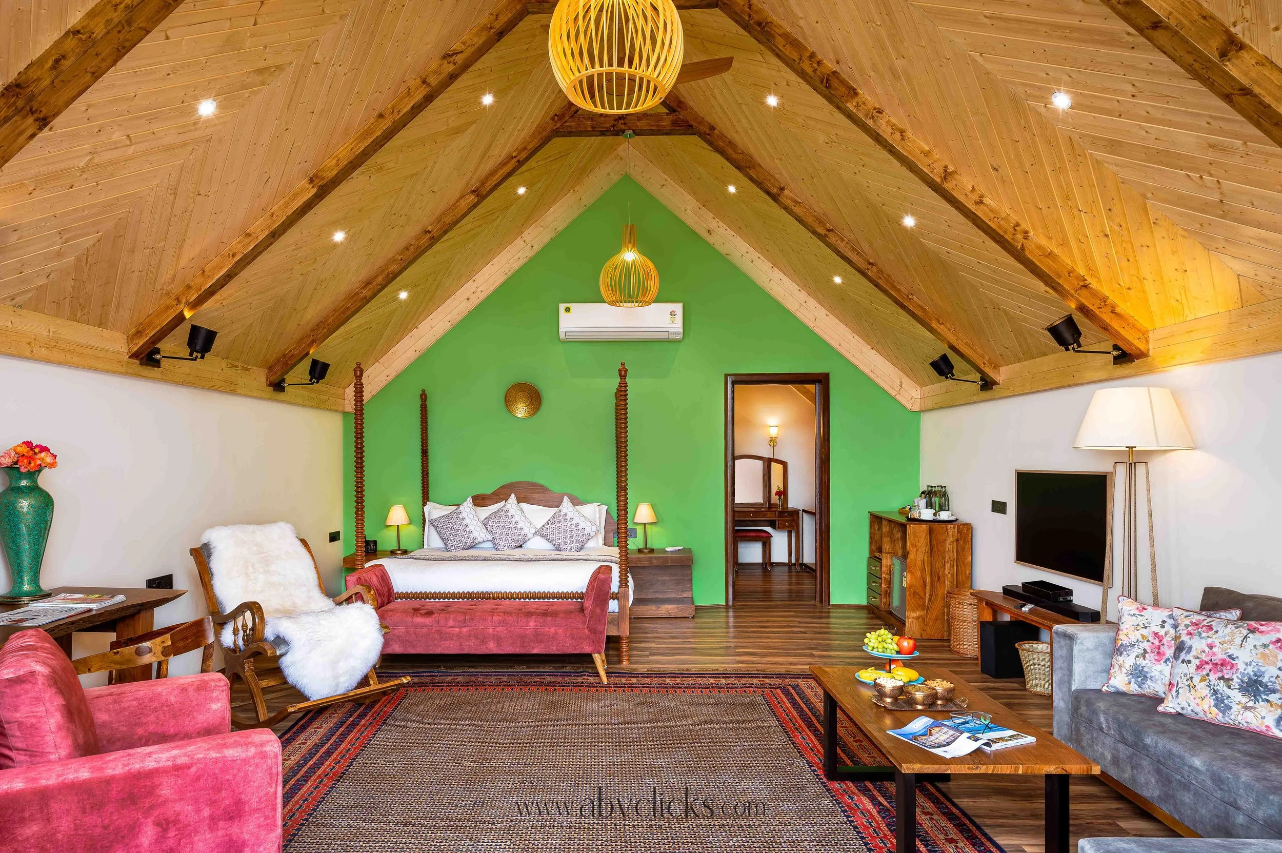 A living room with a vaulted wooden ceiling, green accent wall, bed, pink velvet armchair with white faux fur throw, wooden coffee table with snacks, floral cushion on gray sofa, television, and decorative lighting fixtures.