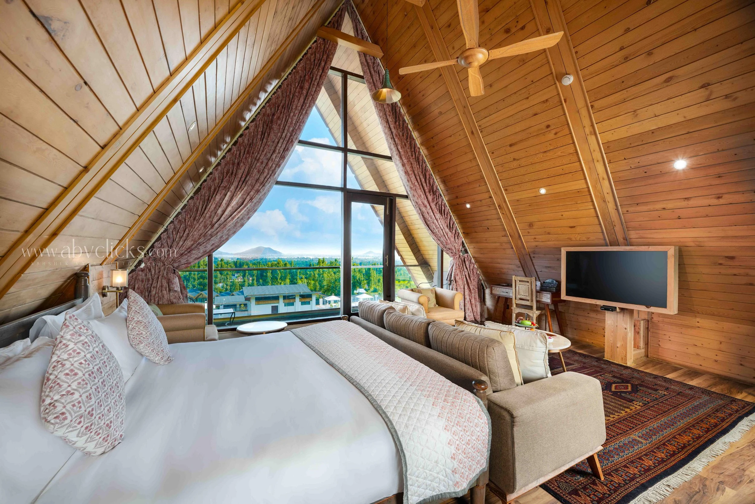 Interior of a wooden ceiling cabin bedroom with a large bed, a sitting area, and a large window showing a view of trees and mountains.