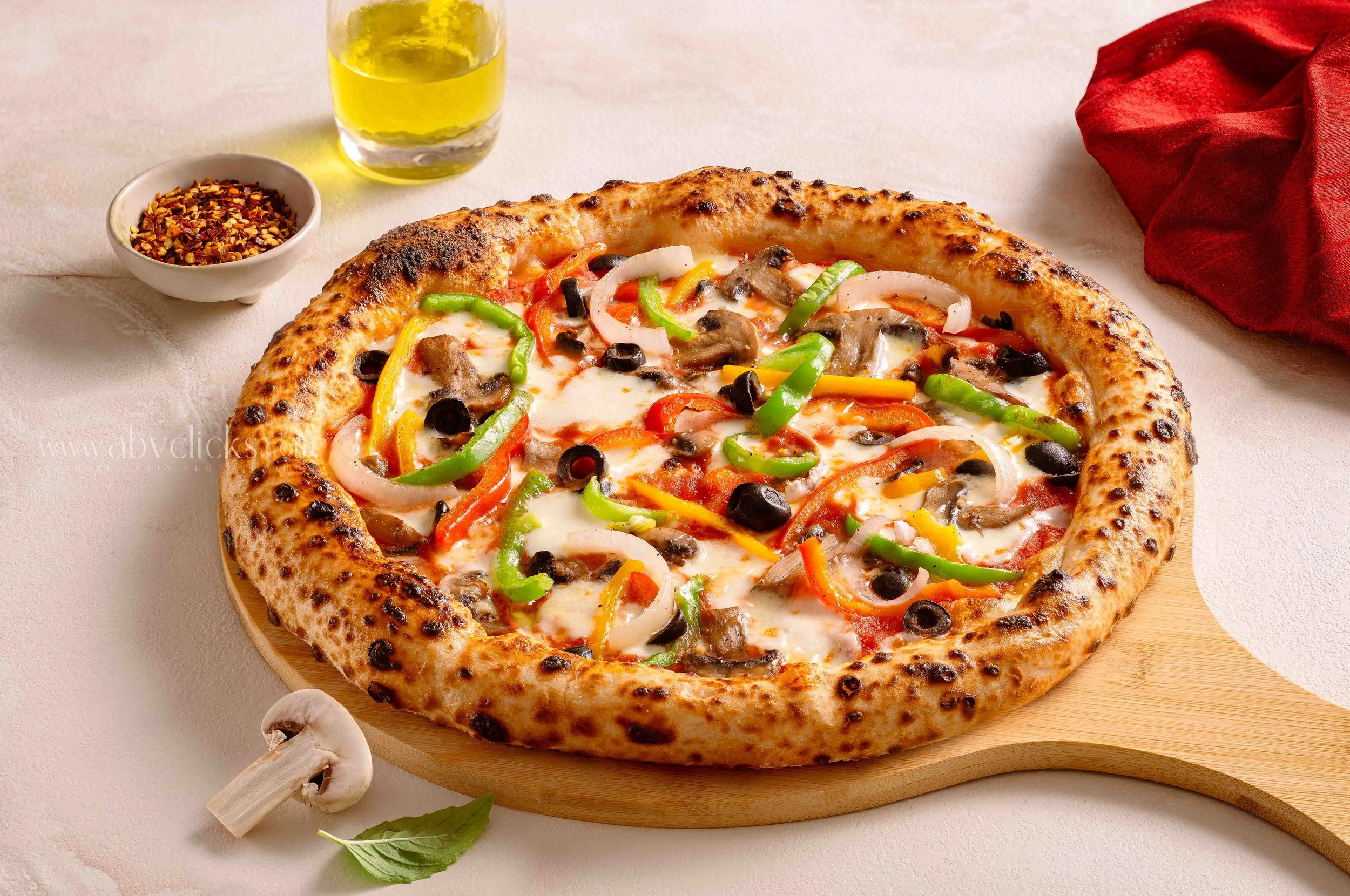 A pizza with cheese, peppers, onions, mushrooms, and black olives on a wooden serving board, with a bowl of red pepper flakes, a glass of yellow liquid, and a red cloth napkin nearby.