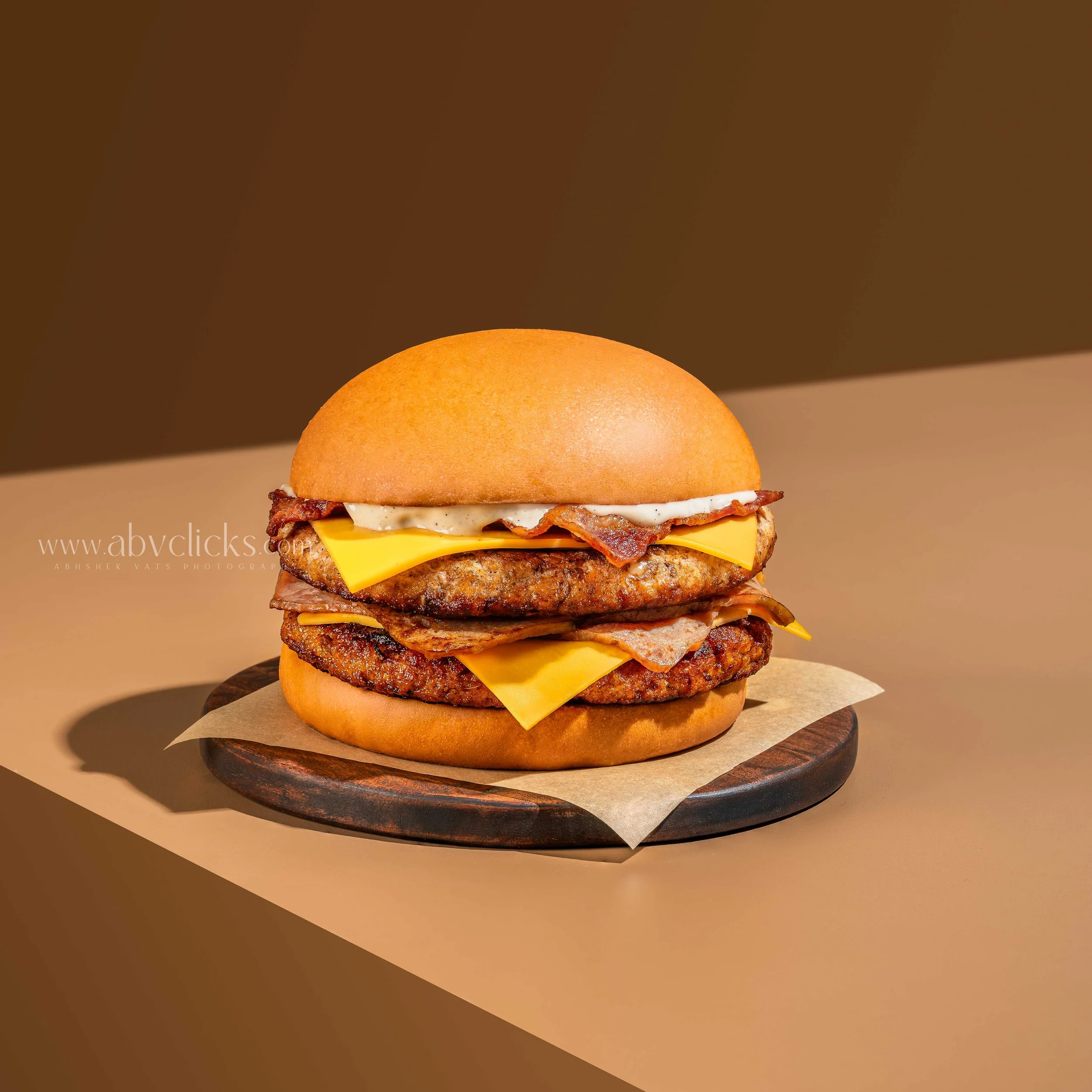 Close-up of a burger with bacon and cheese on a sesame seed bun, placed on a dark wooden plate with parchment paper beneath.