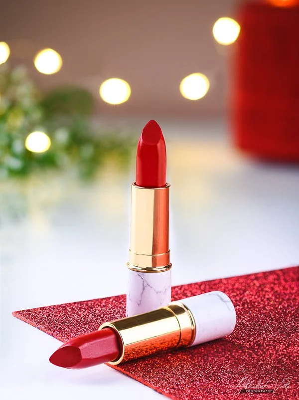 Two red lipsticks, one standing upright and the other lying down on a red glittery surface, with blurred holiday lights and greenery in the background.