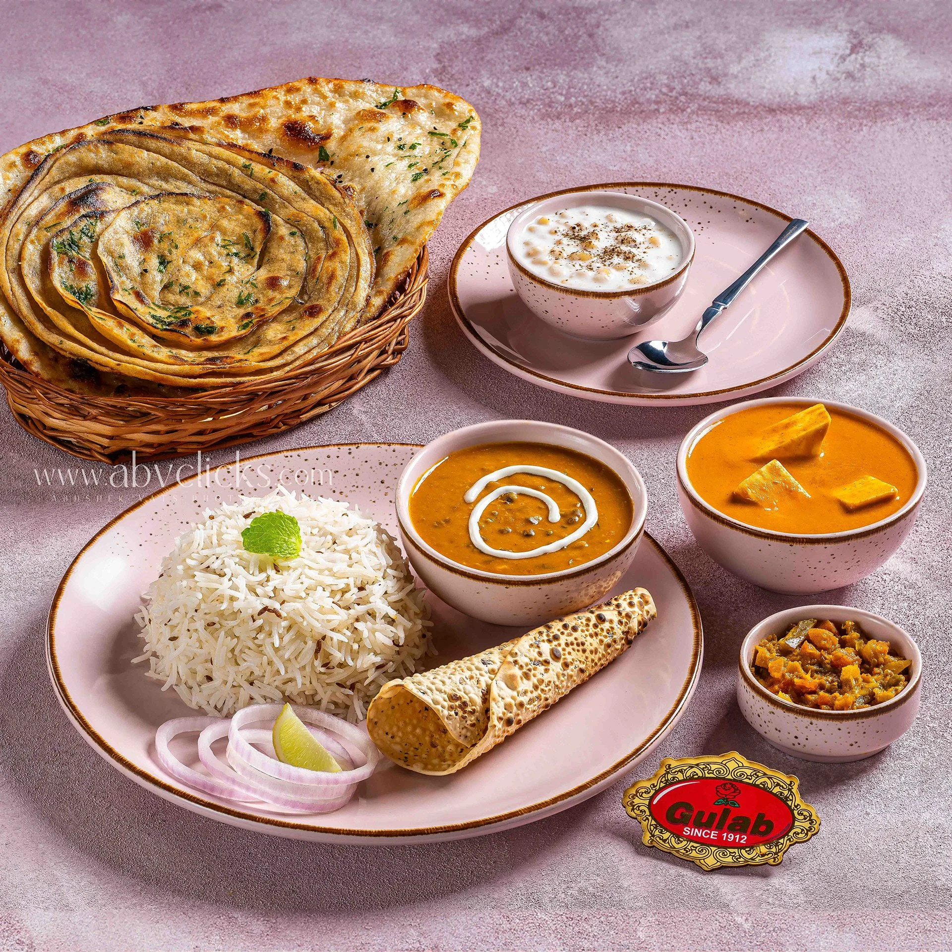 Plate with white rice garnished with a lime wedge and onion slices, bowl of lentil curry topped with sour cream and cilantro, bowl of vegetable curry, piece of flatbread, basket with stuffed flatbread, and small bowls with various Indian dishes.