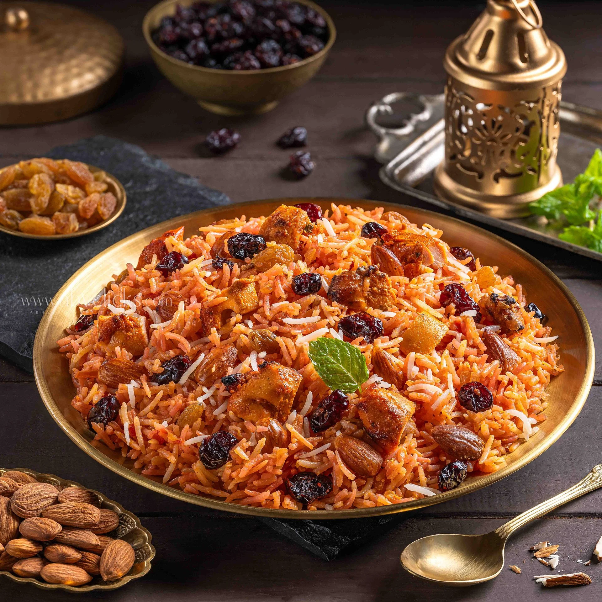 A plate of saffron rice pilaf garnished with dried cranberries and almonds, surrounded by bowls of dried cranberries, almonds, and cardamom pods, with a brass incense holder and a gold-colored spoon on a dark wooden surface.