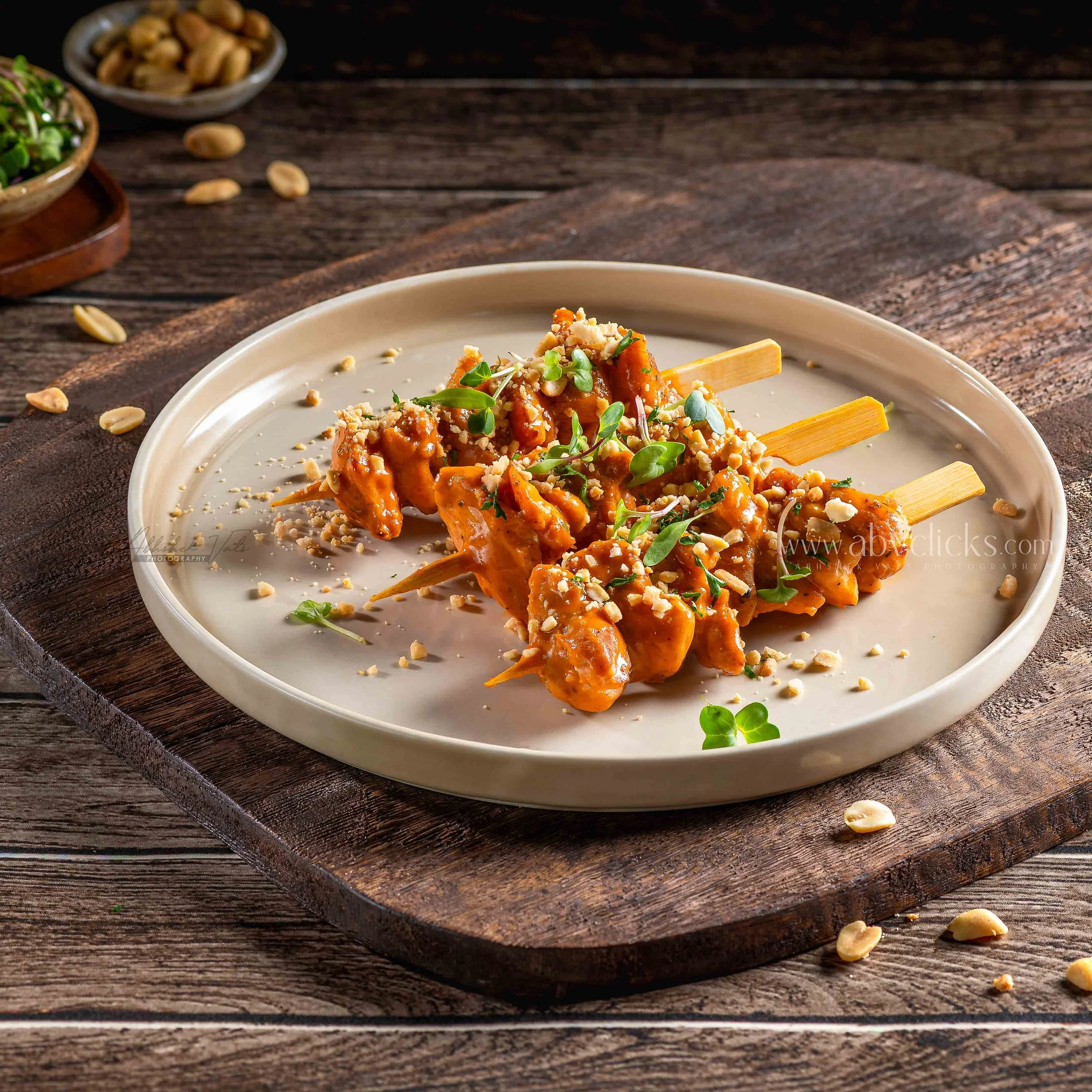 Close-up of grilled chicken skewers on a white plate, garnished with chopped nuts and microgreens, placed on a wooden surface. In the background, there are bowls of nuts and herbs.