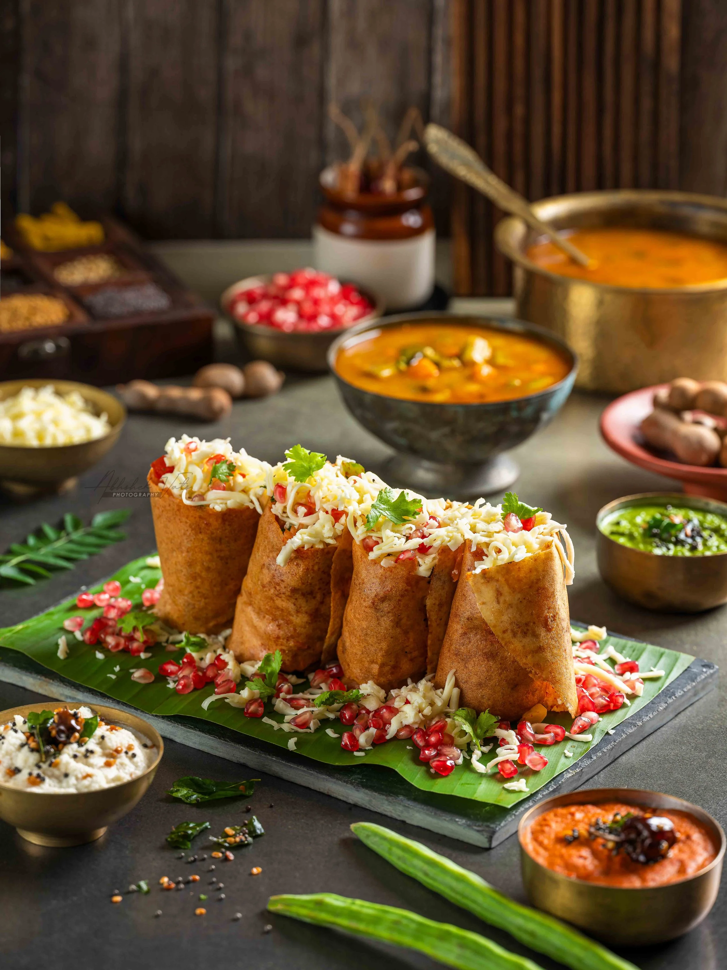 Indian street food featuring several pani puris topped with shredded cheese and garnished with pomegranate seeds, surrounded by small bowls of various colorful chutneys and side dishes on a black slate platter with green leaves and scattered seeds.