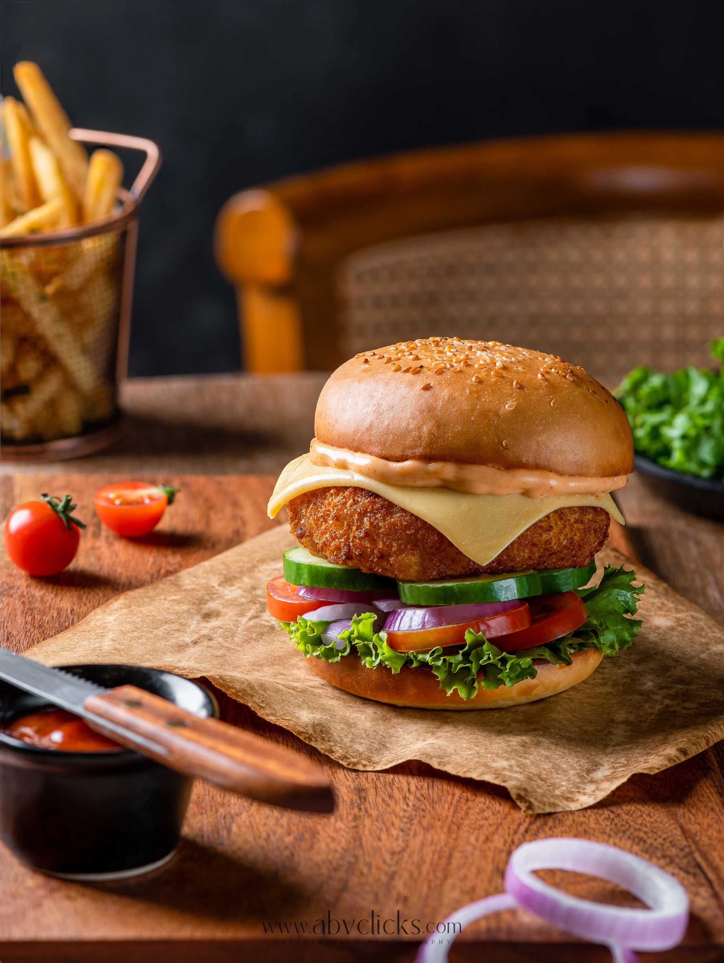 A close-up of a crispy chicken sandwich with lettuce, tomato, sliced onions, cheese, and a sesame seed bun on a piece of brown paper. In the background, there are cherry tomatoes, a bowl of French fries, and a wooden chair.