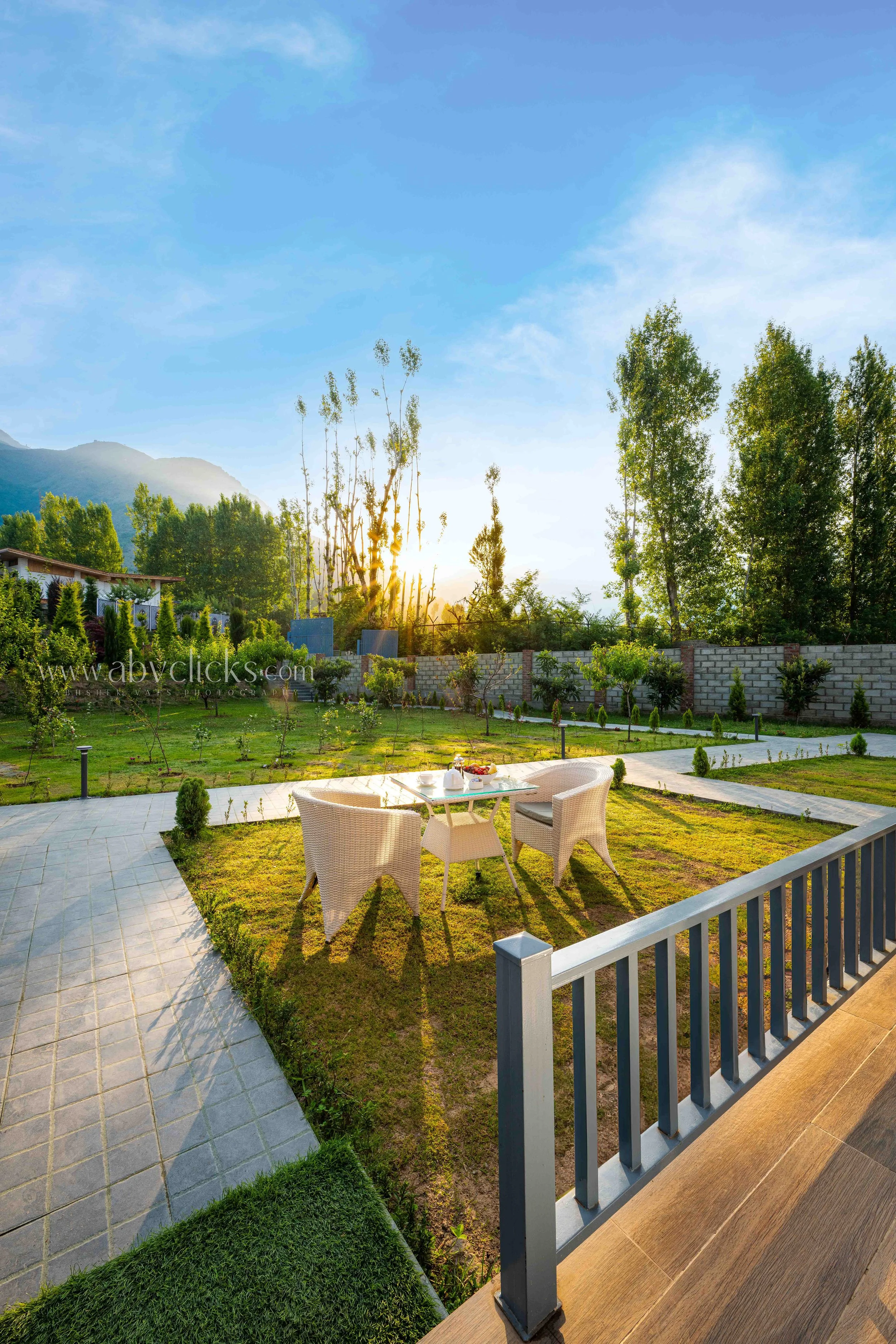 Sunset view from a balcony overlooking a garden with a table and chairs, trees, a stone pathway, and a mountain in the background.