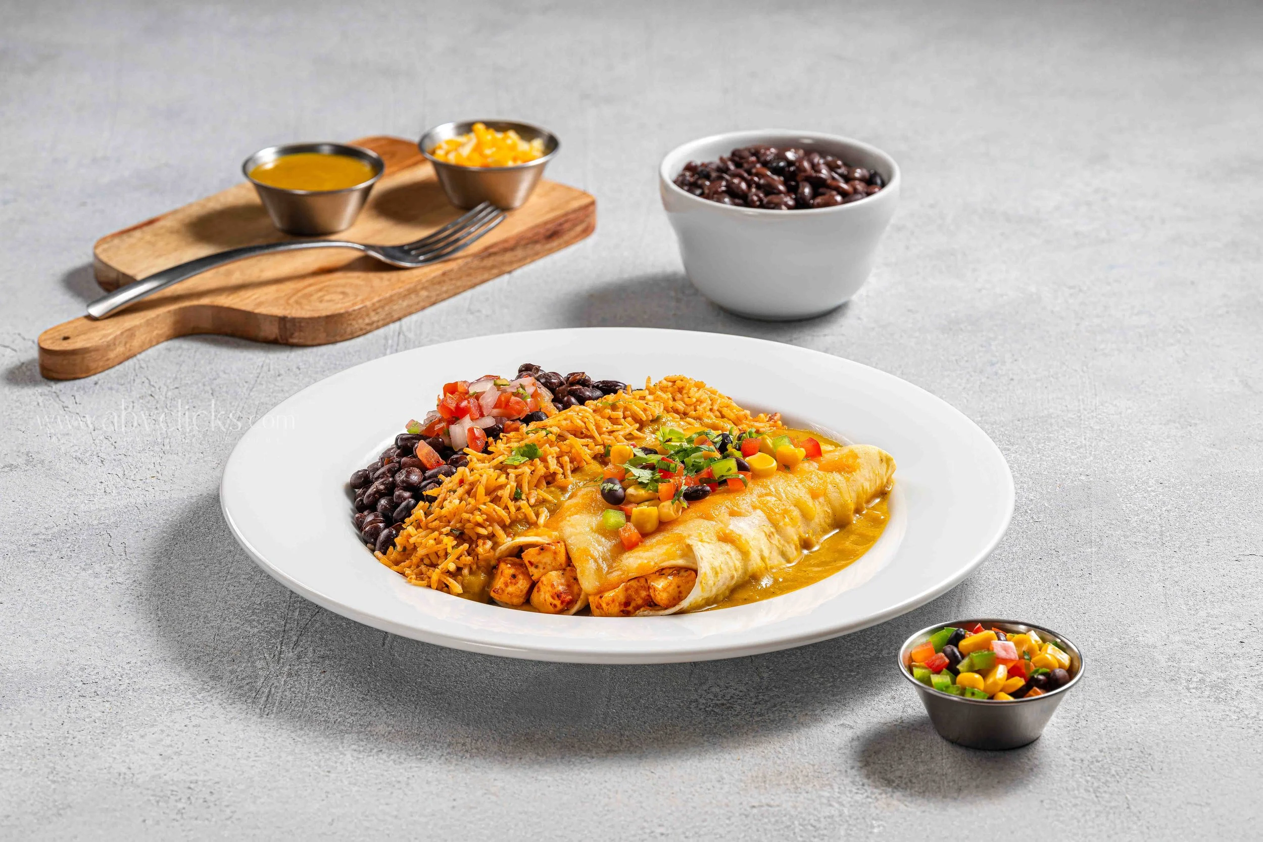 Plate of Mexican food with rice, black beans, chicken enchilada topped with cheese, and colorful vegetable garnish, with side bowls of black beans and salsa in the background.