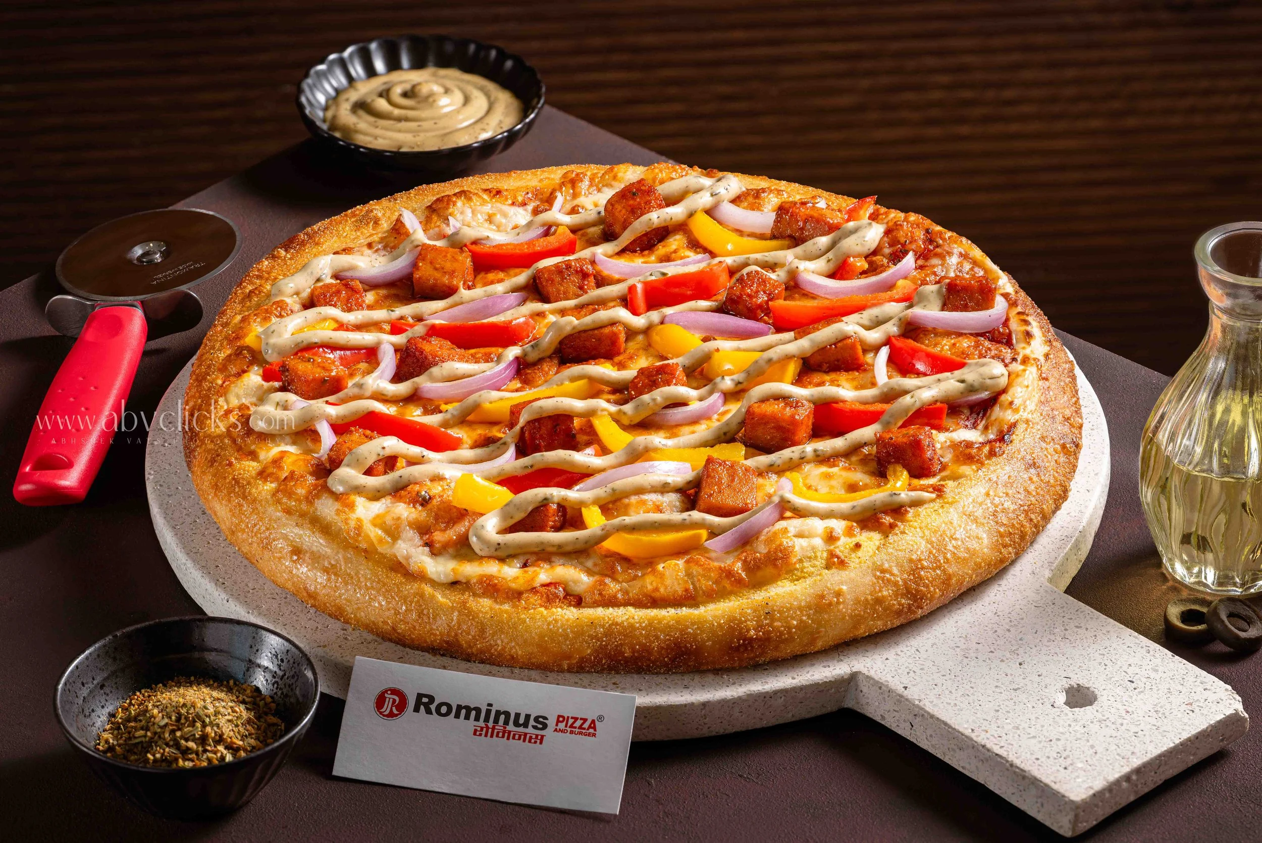 A whole pizza topped with sausage, red and yellow bell peppers, onions, and drizzled with white sauce, on a pizza stone. A small bowl of mixed herbs and a bowl of crushed red pepper flakes are nearby, with a small sign for Rominus Pizza and Burger.