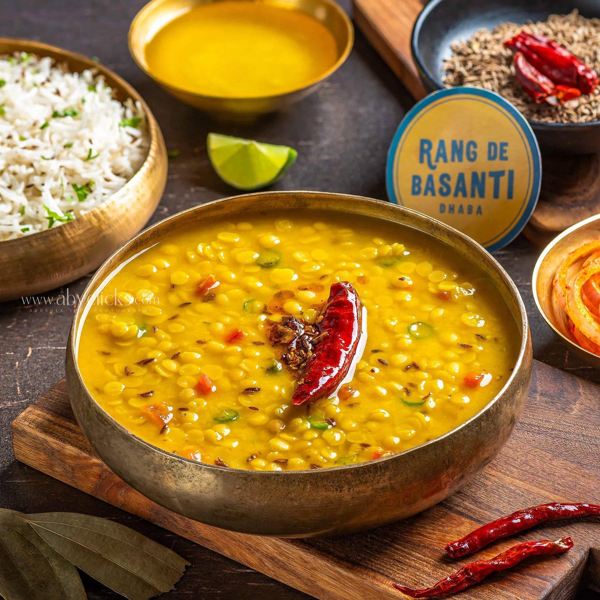 A bowl of yellow lentil soup with a red chili, sitting on a wooden surface with a cutting board and other bowls of food nearby.