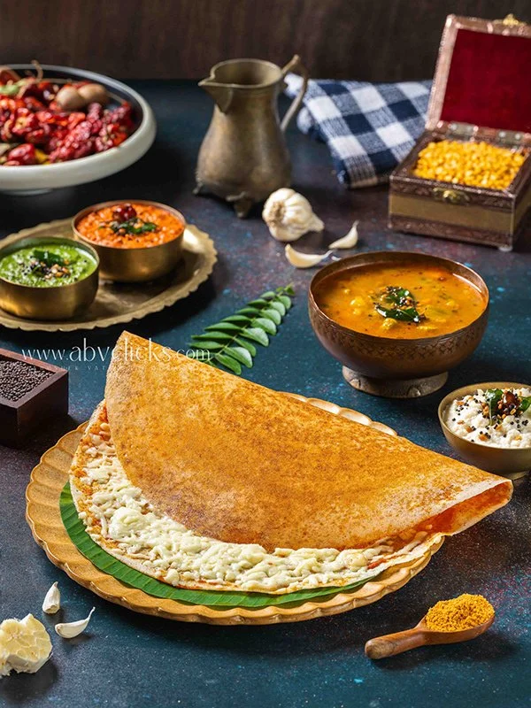 Indian meal with a large stuffed paratha on a green plate, served with various bowls of curries and side dishes, garlic, spices, and a box of popcorn on a dark surface.