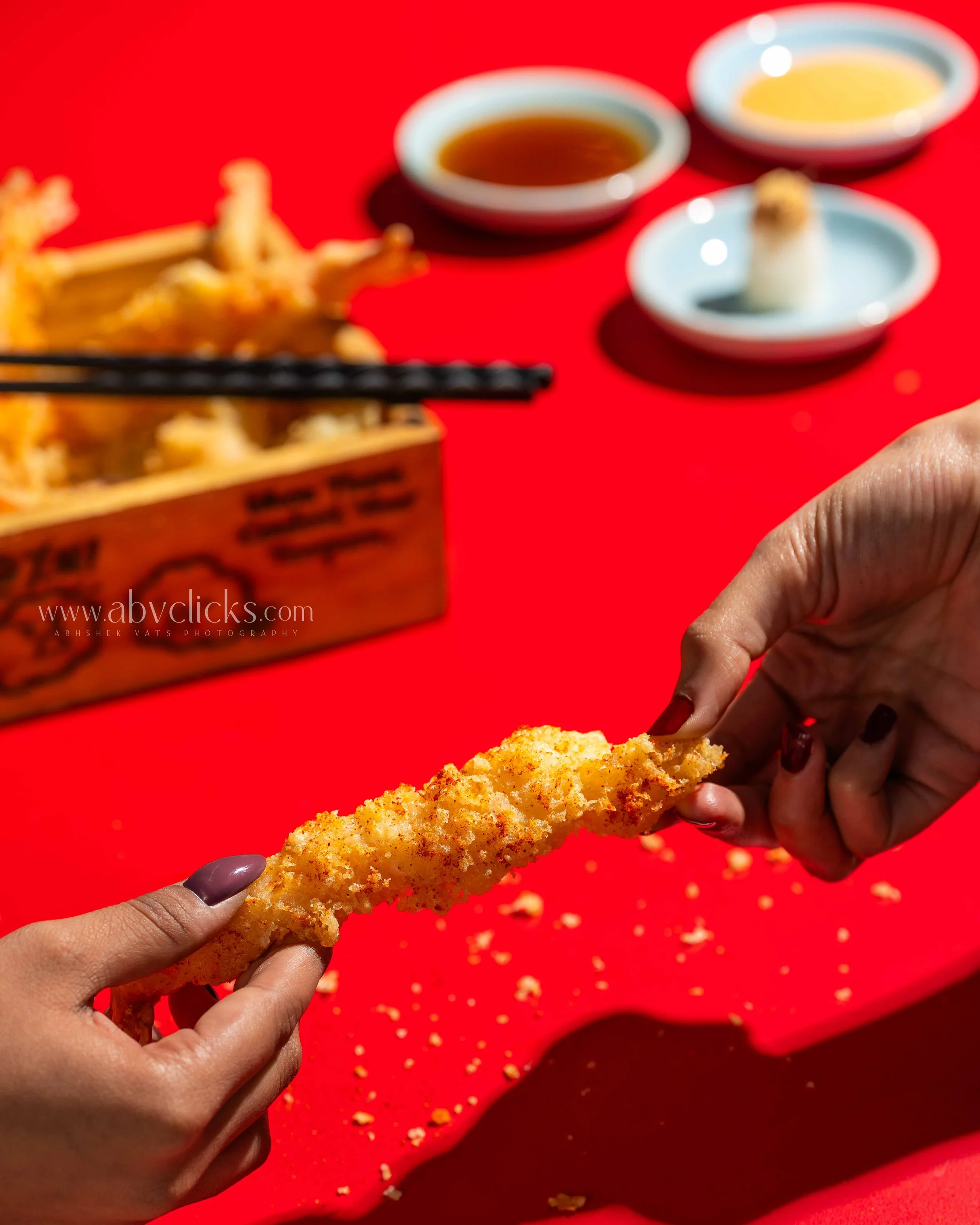 Person holding a piece of fried chicken with a bright red background, two small bowls of dipping sauces, and a box of fried chicken in the background.