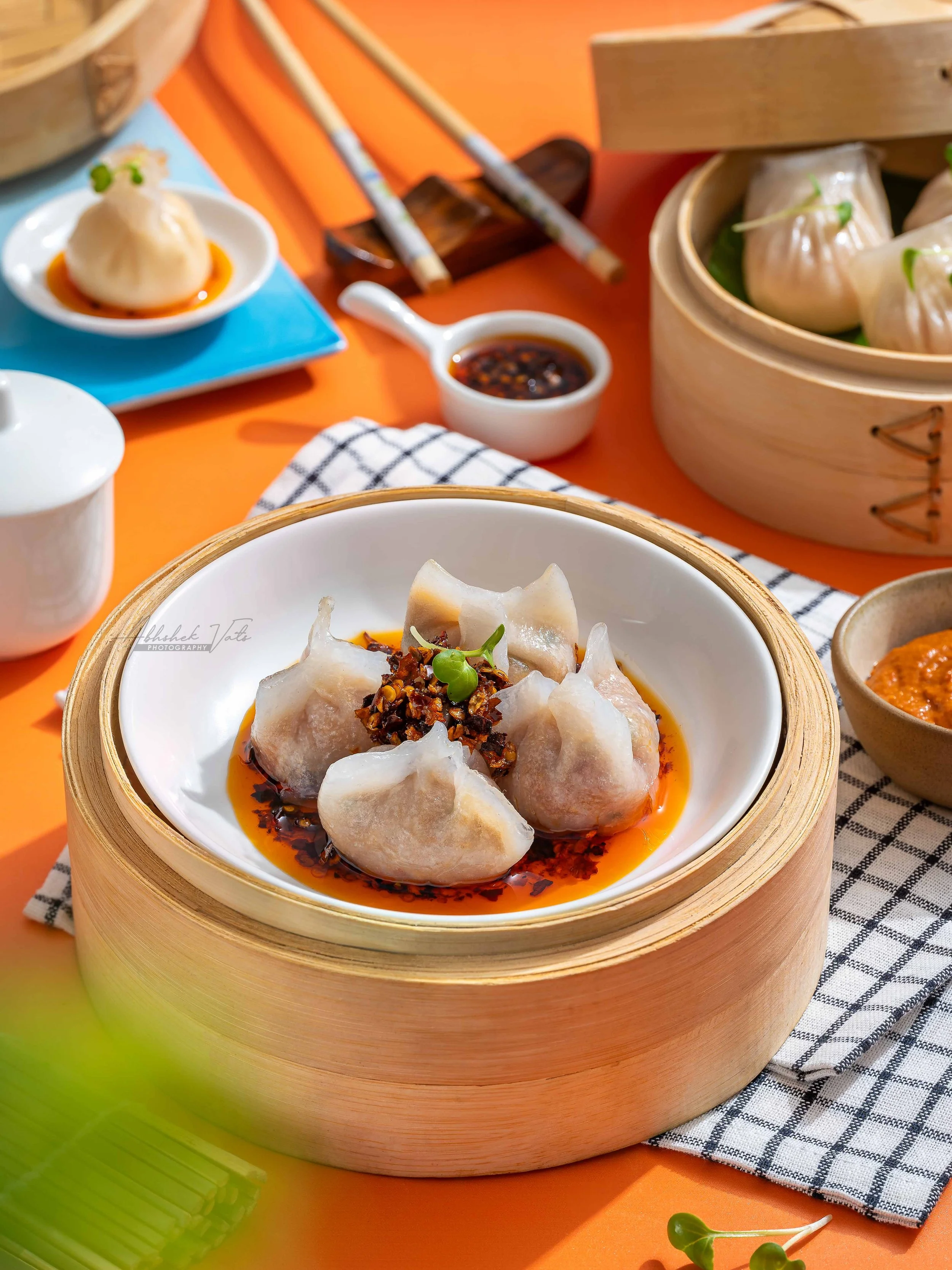 Steamed dumplings garnished with chopped peanuts and green herb, served with soy sauce, on a white plate and wooden steamer basket, with chopsticks and a checkered napkin on an orange table.