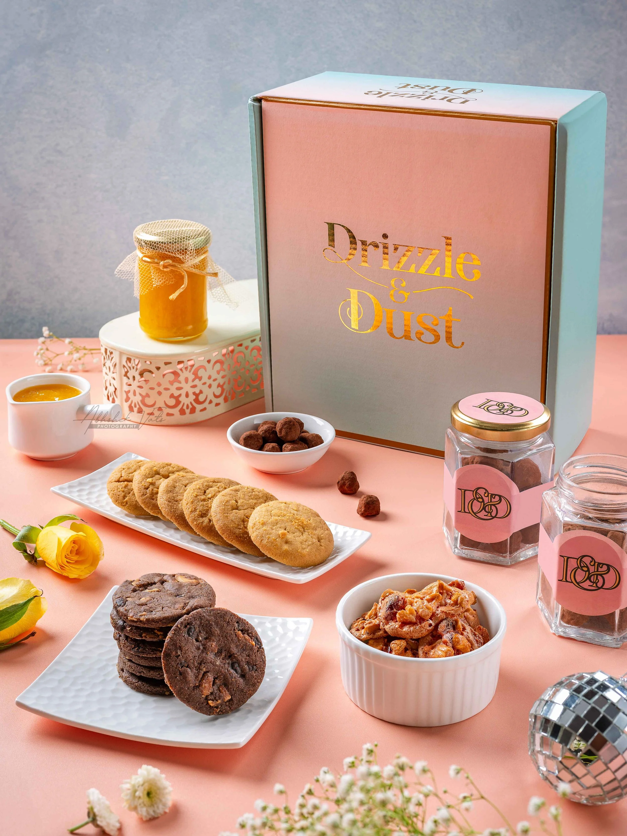 A pink table with an assortment of cookies, chocolates, and bowls of snacks, along with a pastel gift box labeled "Drizzle & Dust," decorative jars, and flowers in the foreground.