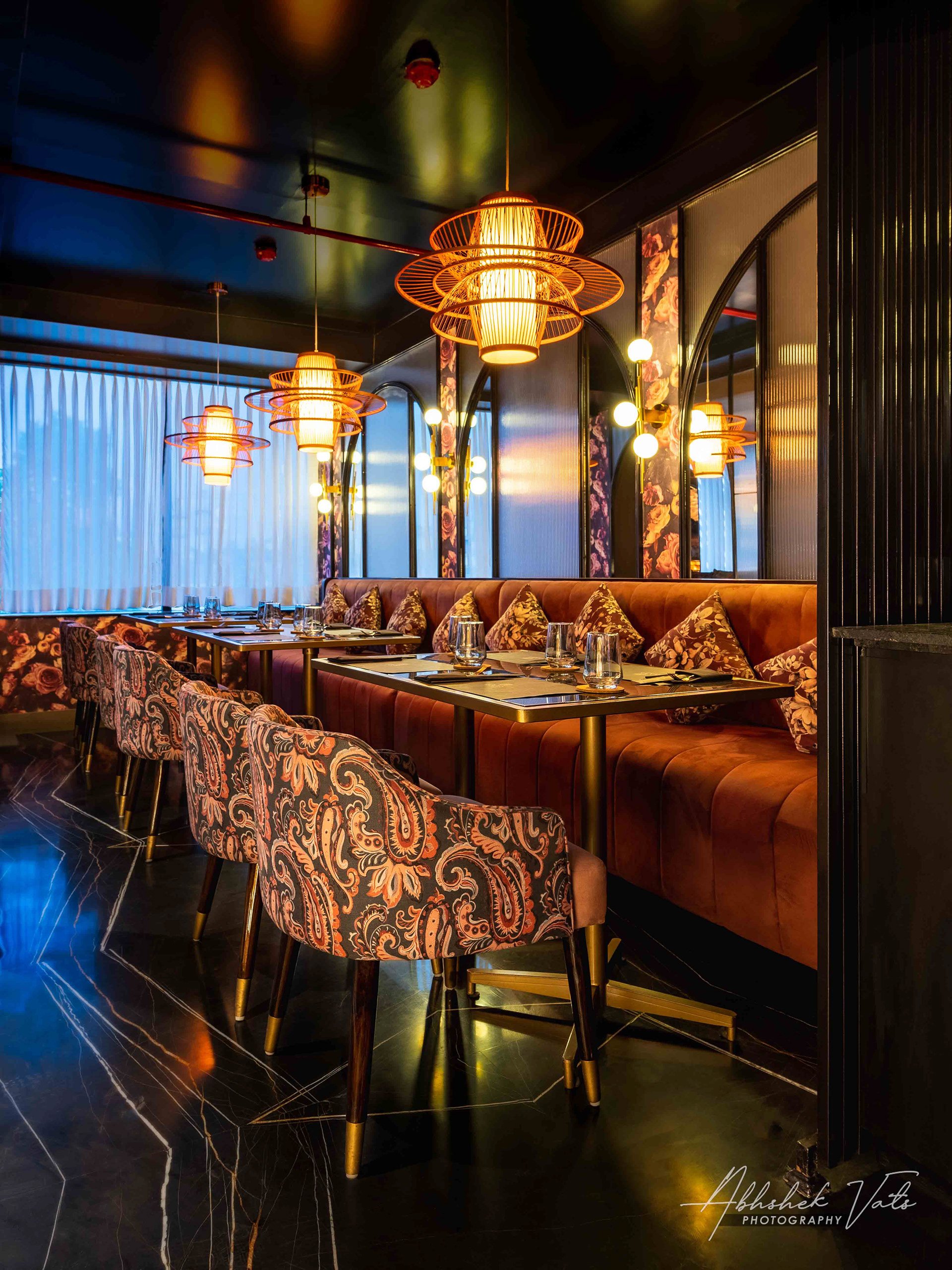 Elegant restaurant interior with a dark ceiling and walls, featuring warm golden pendant lights and wall-mounted lamps. The room has a long, plush, floral-patterned sofa with decorative pillows, and a table set with glasses and place mats. The floor 