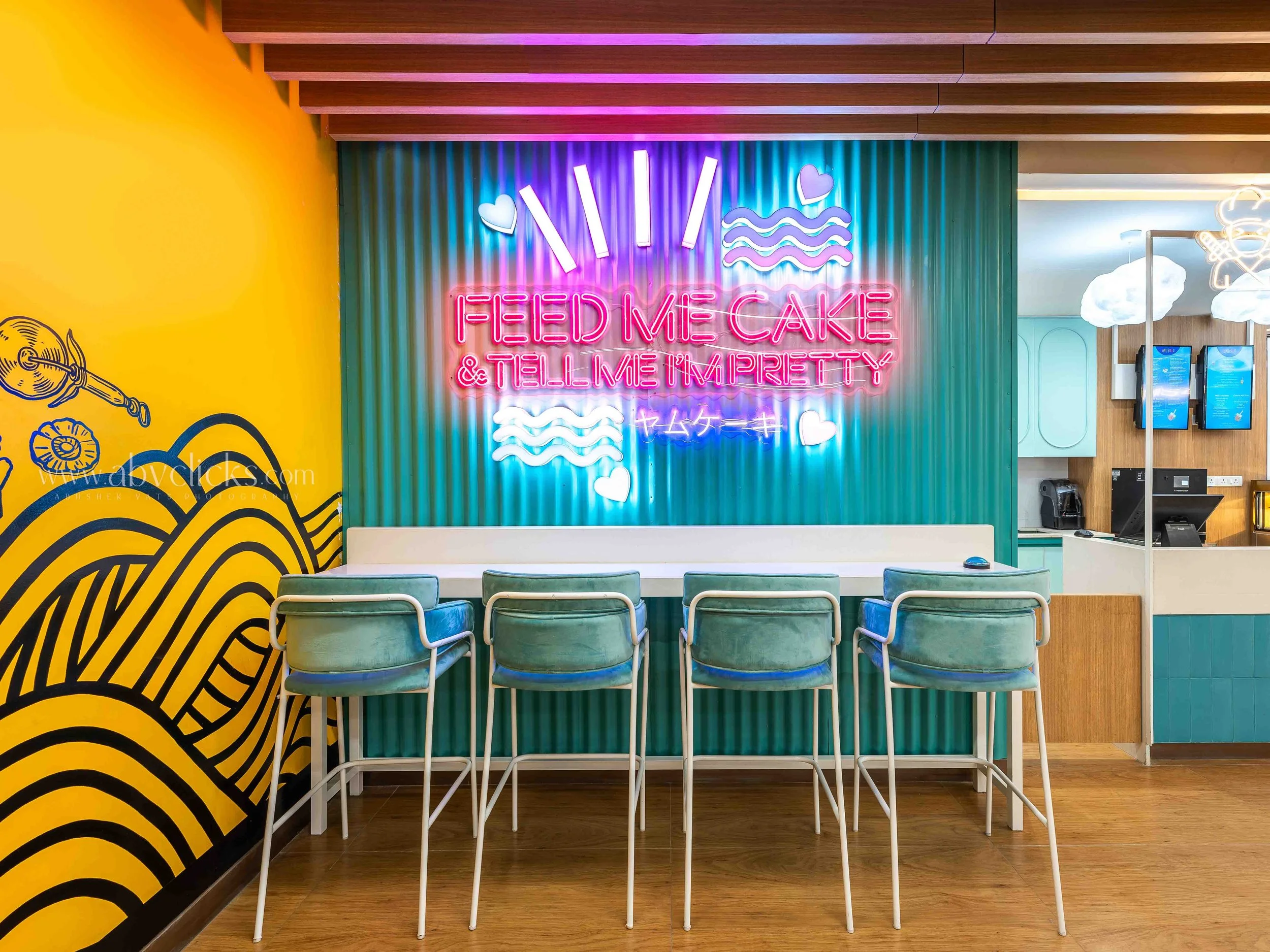 Colorful fast food restaurant interior featuring a neon sign that says 'Feed Me Cake & Tell Me I'm Pretty' on a teal corrugated wall. In front, there are six green chairs with white frames facing the wall, and part of a yellow wall with black wave pa
