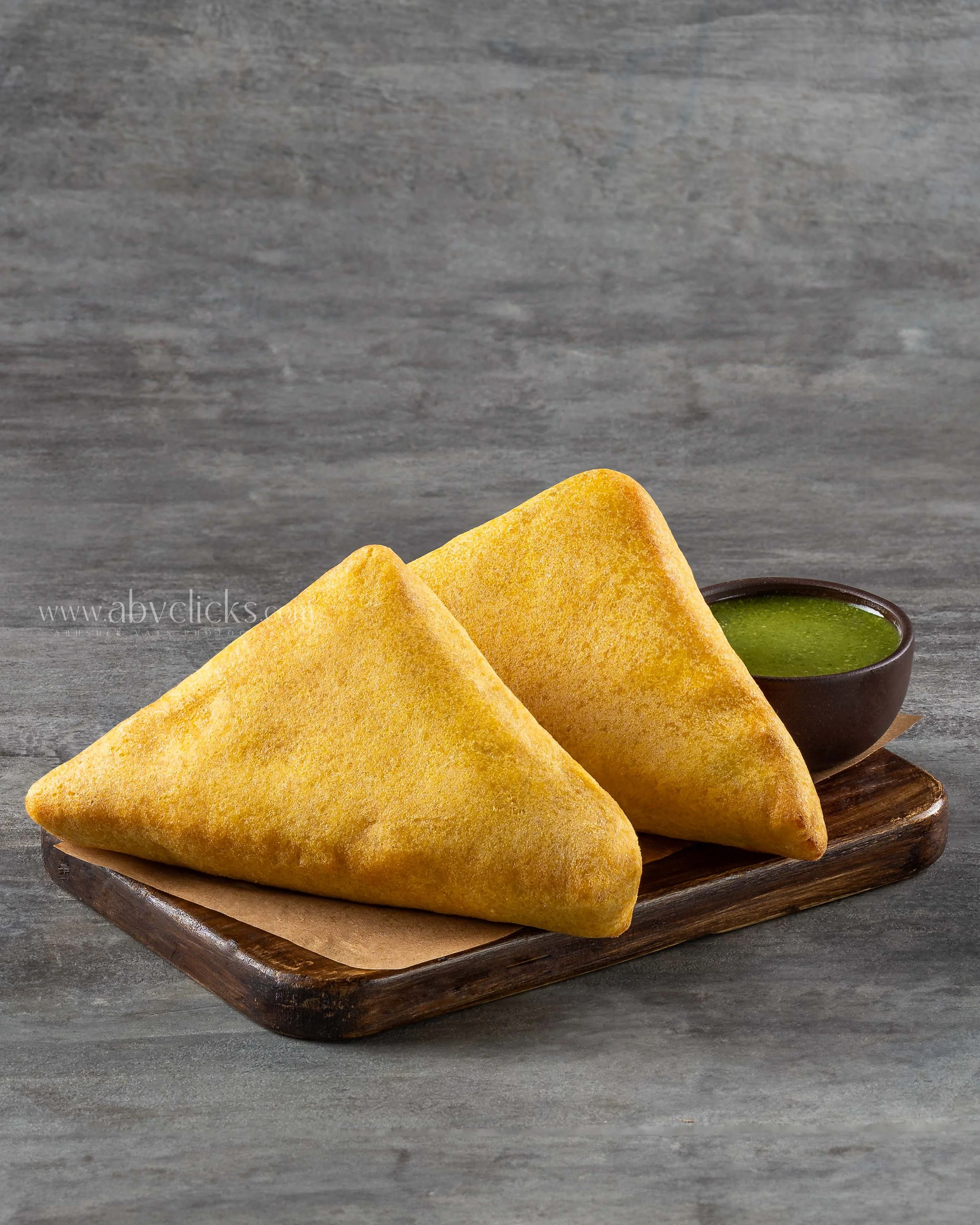 Three golden fried samosas on a wooden serving platter with a small bowl of green cilantro chutney on the side.