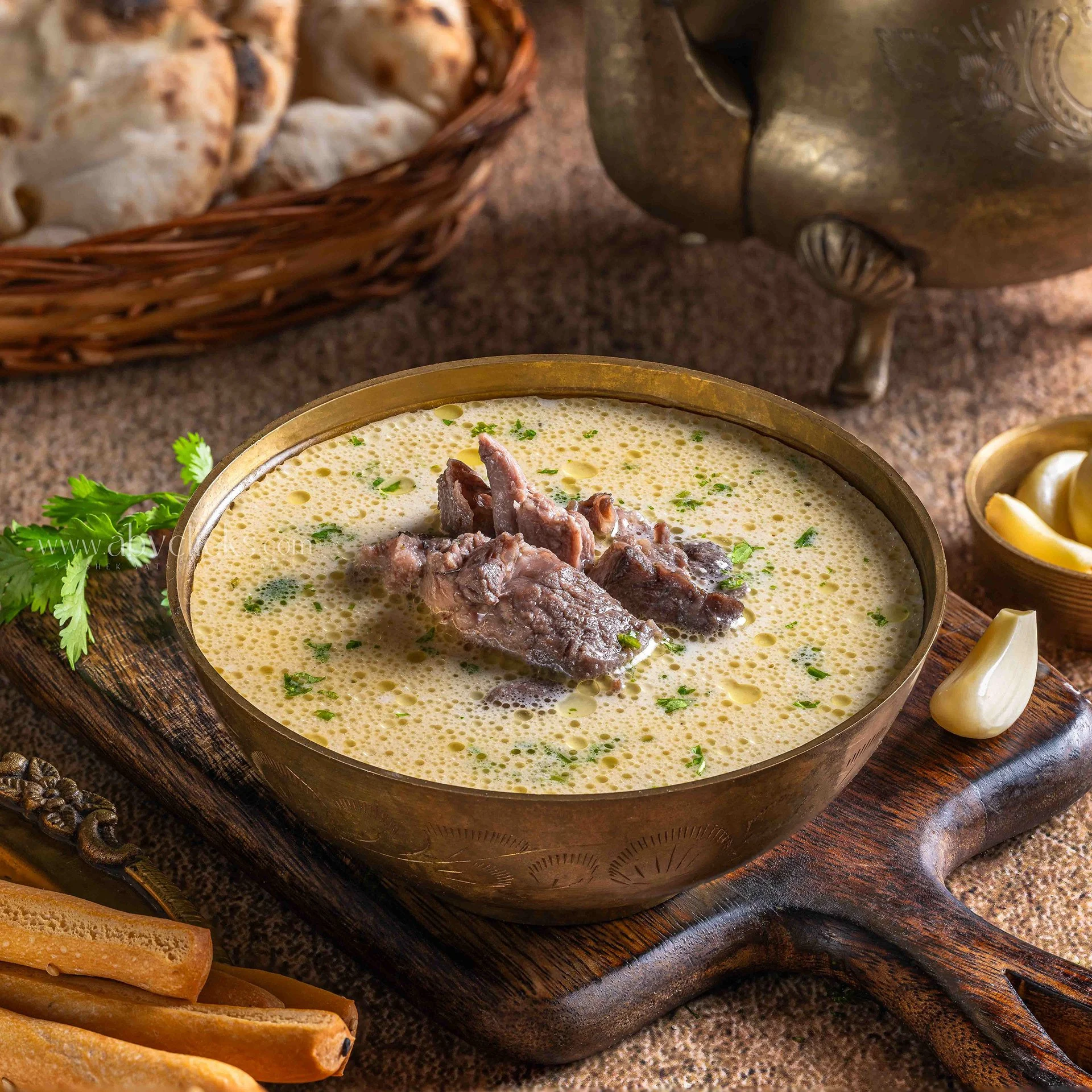 A bowl of Indian curry with pieces of meat garnished with herbs, placed on a wooden surface with a sprig of cilantro nearby. There are also some breadsticks and a golden goblet in the background.