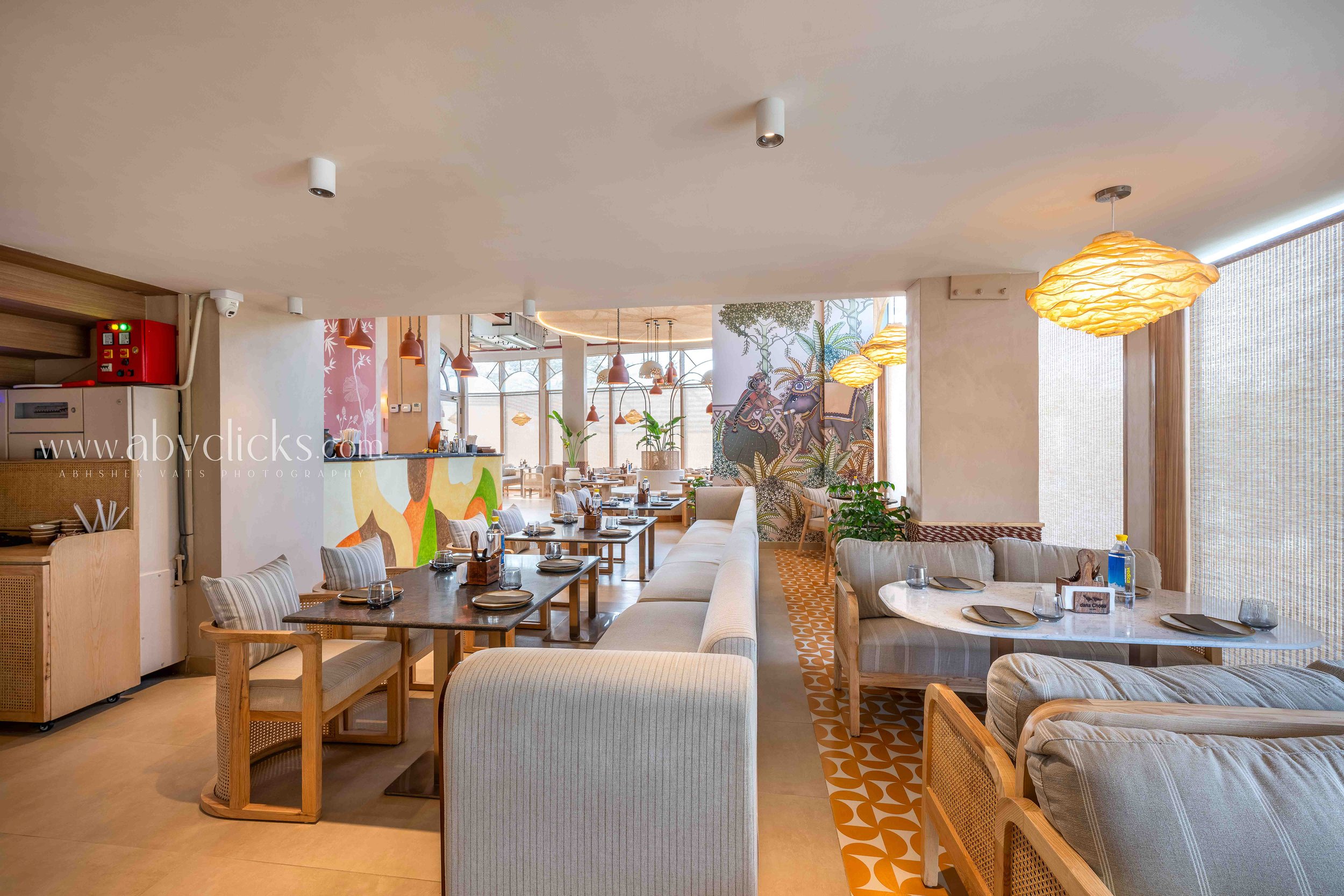 Interior of a bright restaurant with wooden furniture, beige and striped upholstered chairs, decorated with colorful mural wall art, hanging pendant lights, and large windows with sheer curtains.