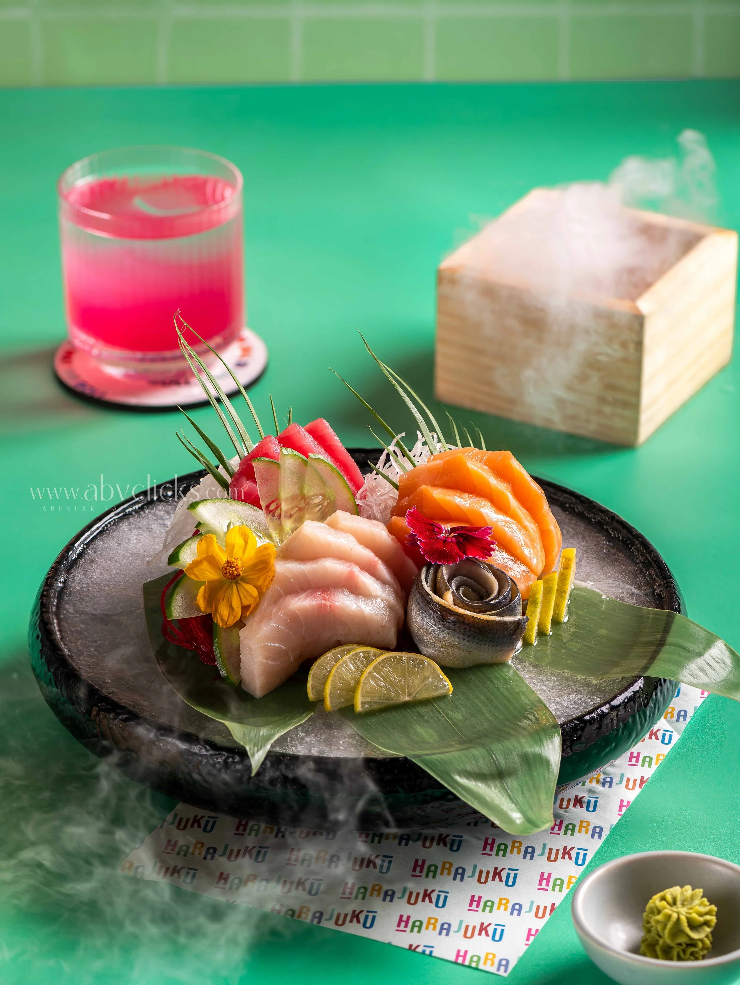Plate of assorted sashimi with lemon slices and edible flowers, glass of pink liquid drink, wooden box, and small bowl of wasabi on colorful paper with letters