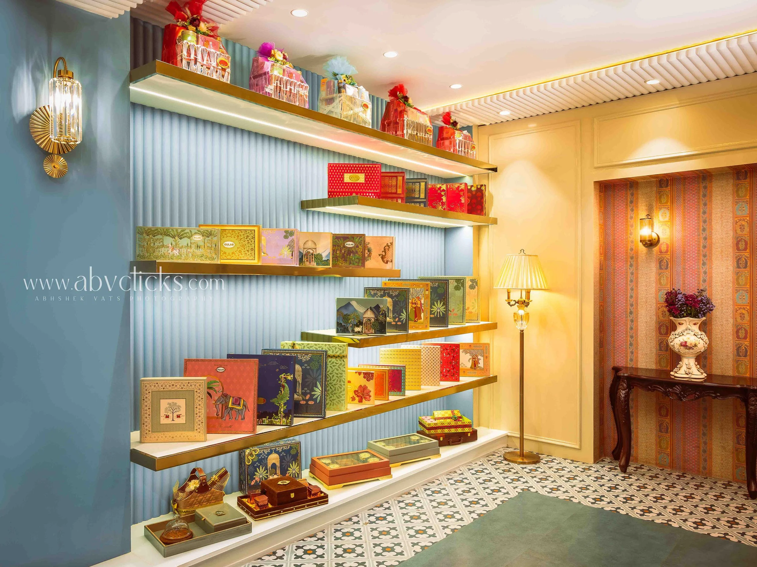 Display of decorative gift boxes and trinkets on white shelves against a blue wall, with patterned tile flooring and a vintage-style lampshade on a table nearby.