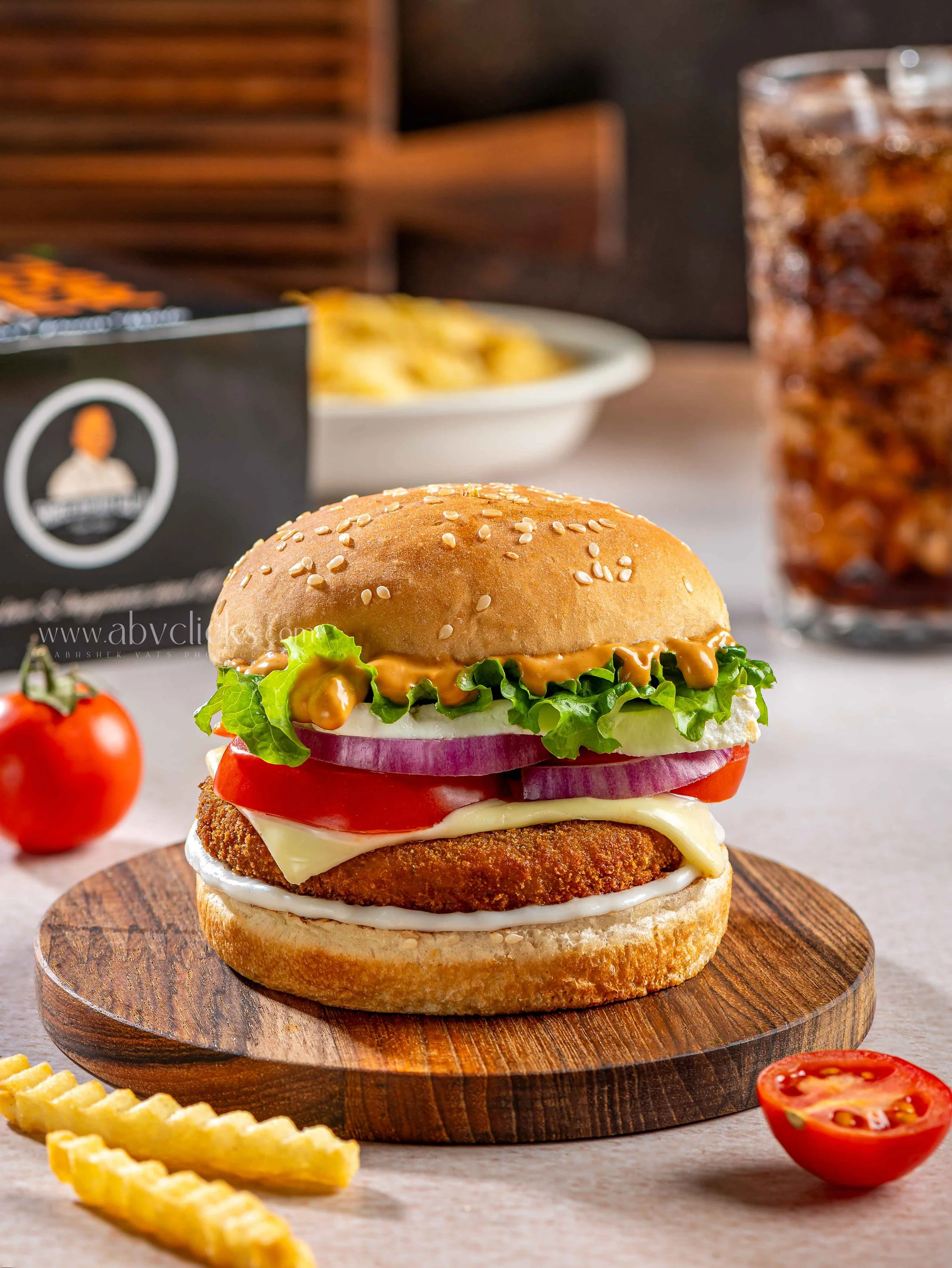 A close-up of a burger with a sesame seed bun, lettuce, tomato, onion, cheese, a fried chicken patty, and sauce, on a round wooden board. In the background, there are potato chips, a soda glass, and a bowl of chips, with some cherry tomatoes and crin
