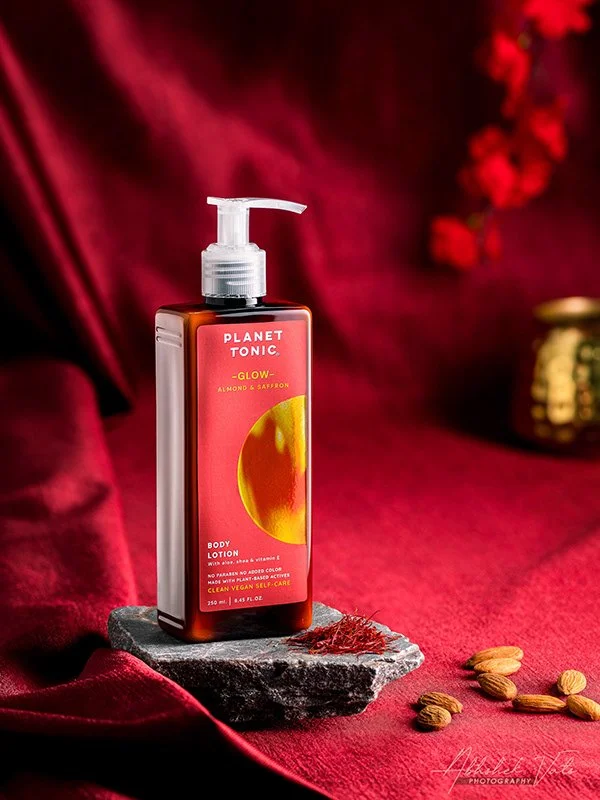 A bottle of Planet Tonic Glow Almond & Saffron body lotion on a piece of gray stone surrounded by almonds, in front of a red velvet background with red flowers and a gold container.