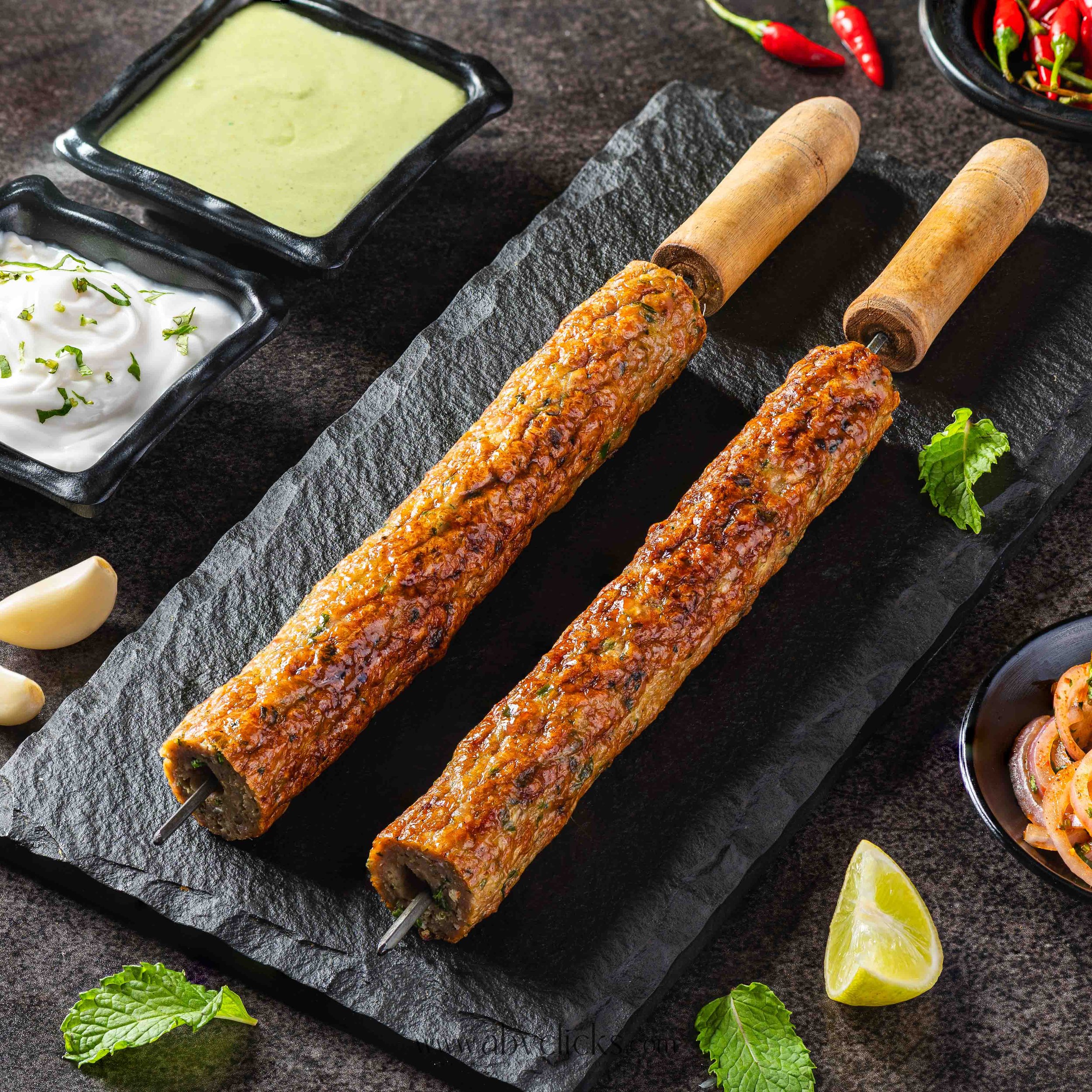 Skewered meat kebabs on a stone plate with lime wedges, herbs, and dipping sauces.