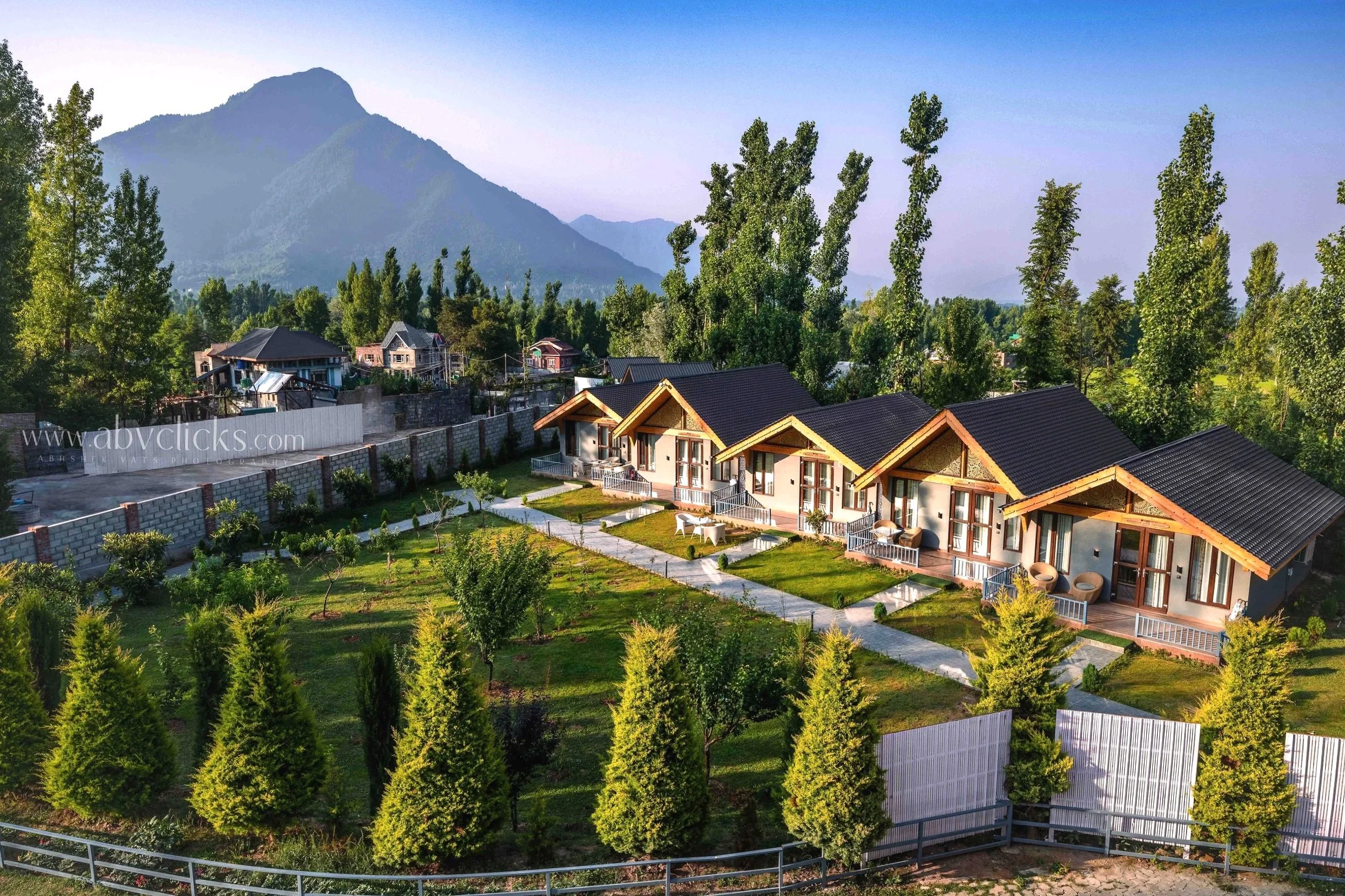 A scenic view of a residential area with modern houses, well-maintained gardens, and tall trees, set against a backdrop of mountains under a clear sky.