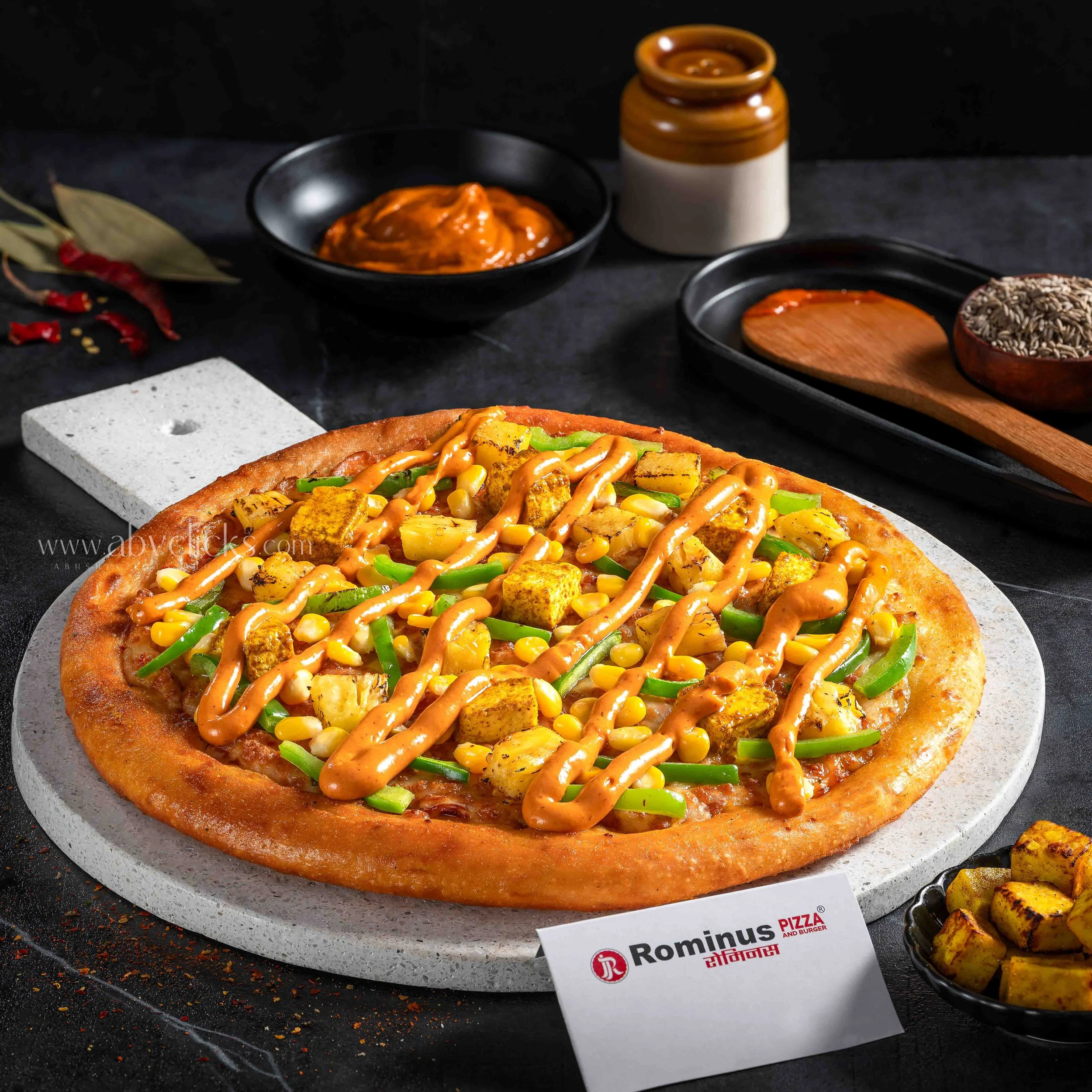 Close-up of a vegan pizza topped with green peppers, corn, grilled tofu, and a drizzle of sauce. The pizza is on a white stand, with a black background and a Rominus Pizza & Burger sign visible. Additional items include a bowl of seeds, a small bowl 