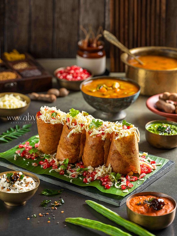 Four Indian-style samosas filled with spices and vegetables, garnished with chopped herbs and pomegranate seeds, served on a banana leaf with various bowls of chutney, yogurt, and curry in a rustic setting.