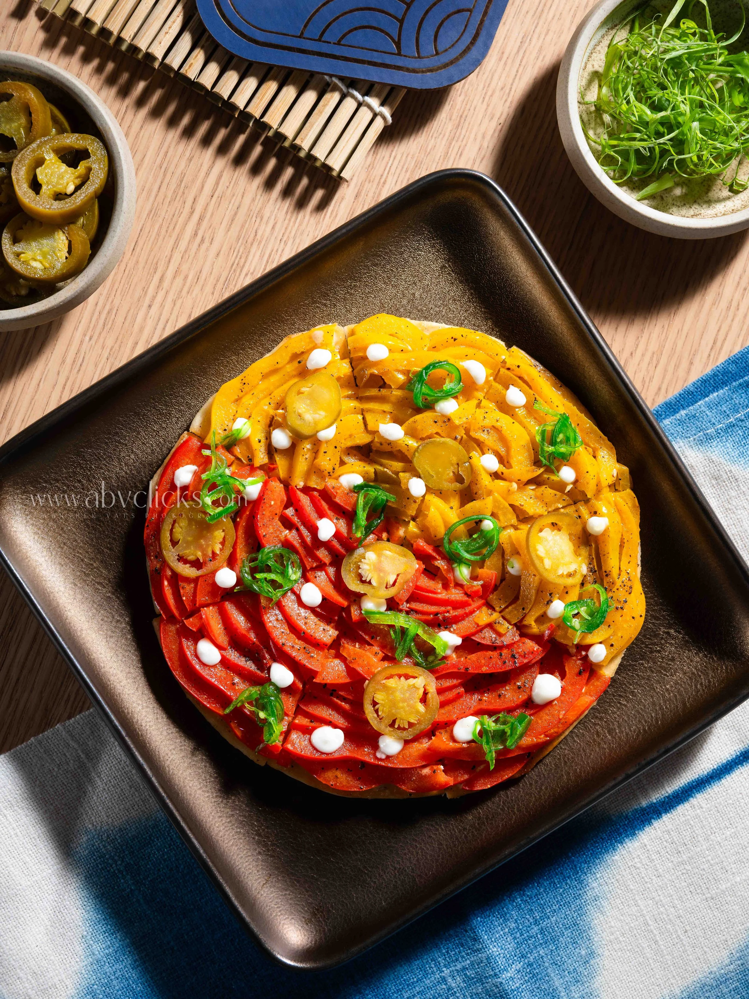 Colored bell pepper rings on a black dish with green onion and dollops of white sauce, on a table with a blue and white tablecloth, alongside small bowls of sliced green peppers and greens, with a bamboo mat and decorative piece in the background.