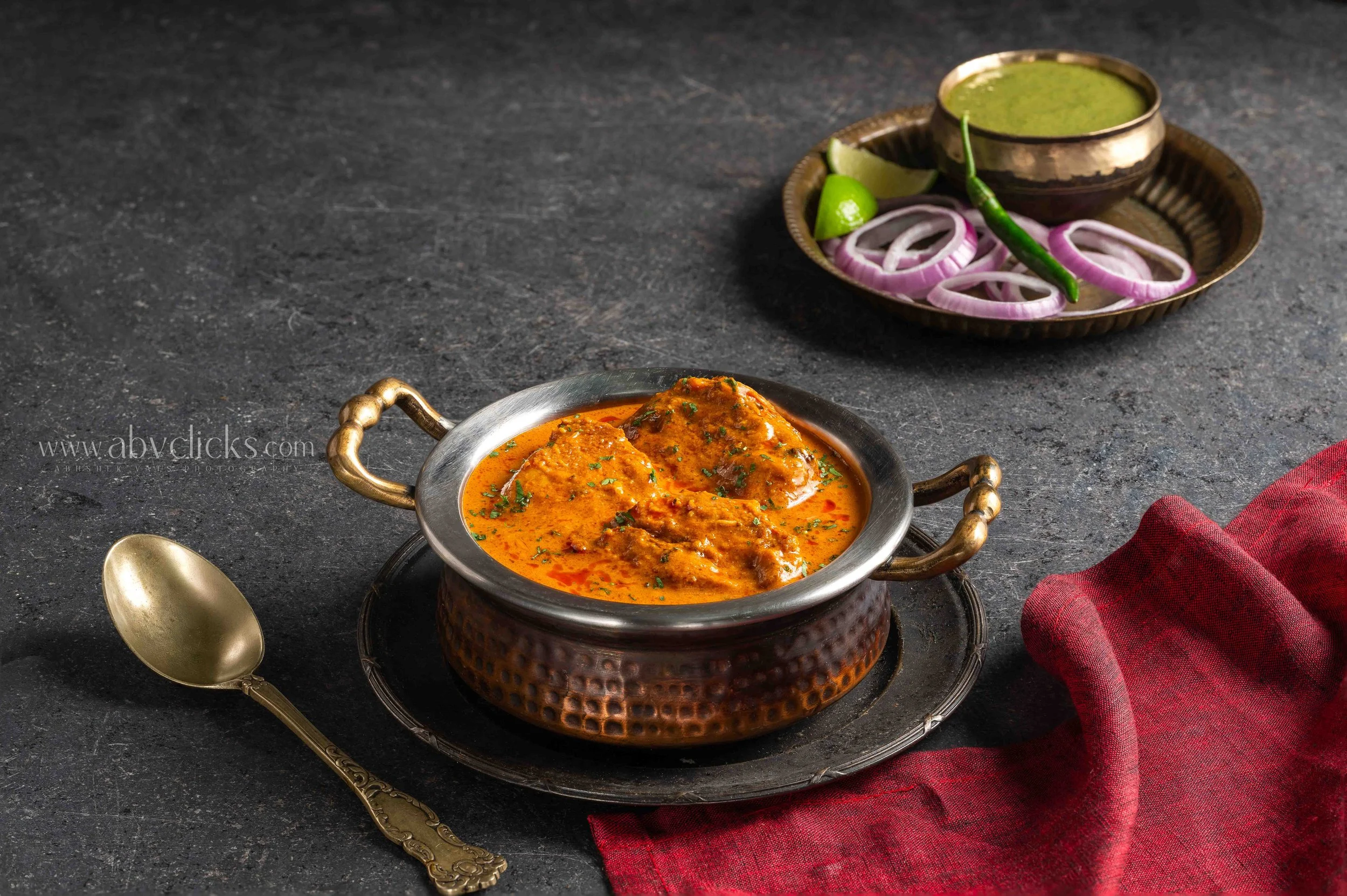 A silver bowl contains Indian chicken curry garnished with herbs, placed on a textured dark surface. In the background, there is a brass plate with sliced onions, lime wedges, and a green chili, along with a small brass bowl of green sauce. A gold sp