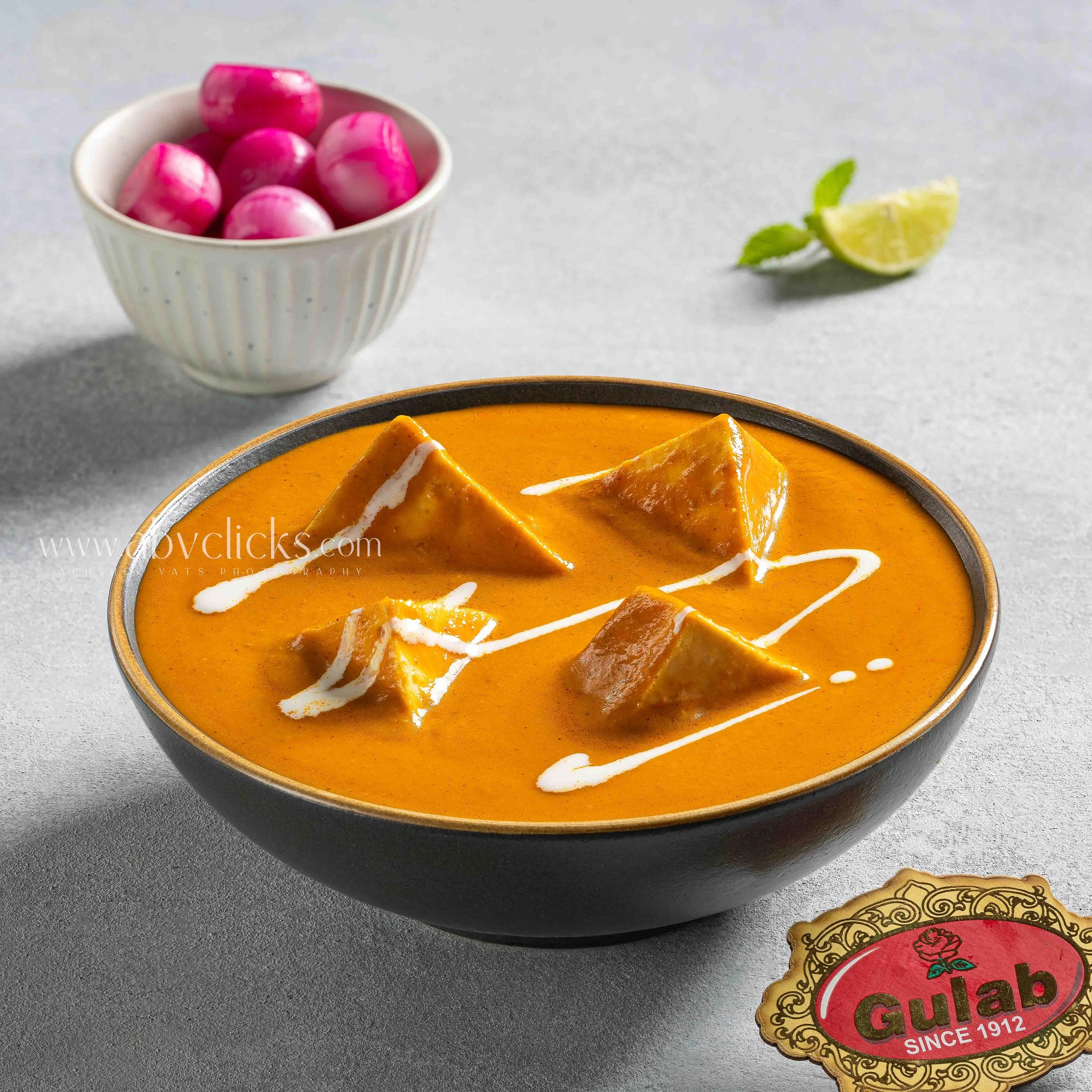 A white ribbed bowl containing pink and white marbles, a slice of lemon with mint leaves, and a portion of a bowl with orange-colored soup garnished with white sauce and triangular chips on a gray surface, a Gula's candy wrapper is partially visible.