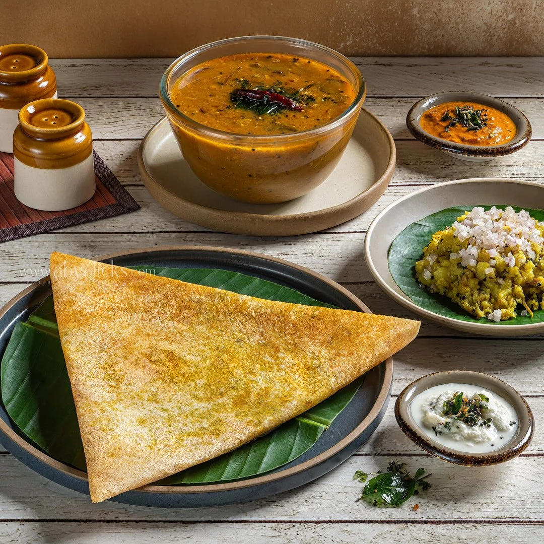 Indian meal with a large crispy dosa, a bowl of sambar, two small bowls of chutney, and a plate of upma, all arranged on a wooden table.