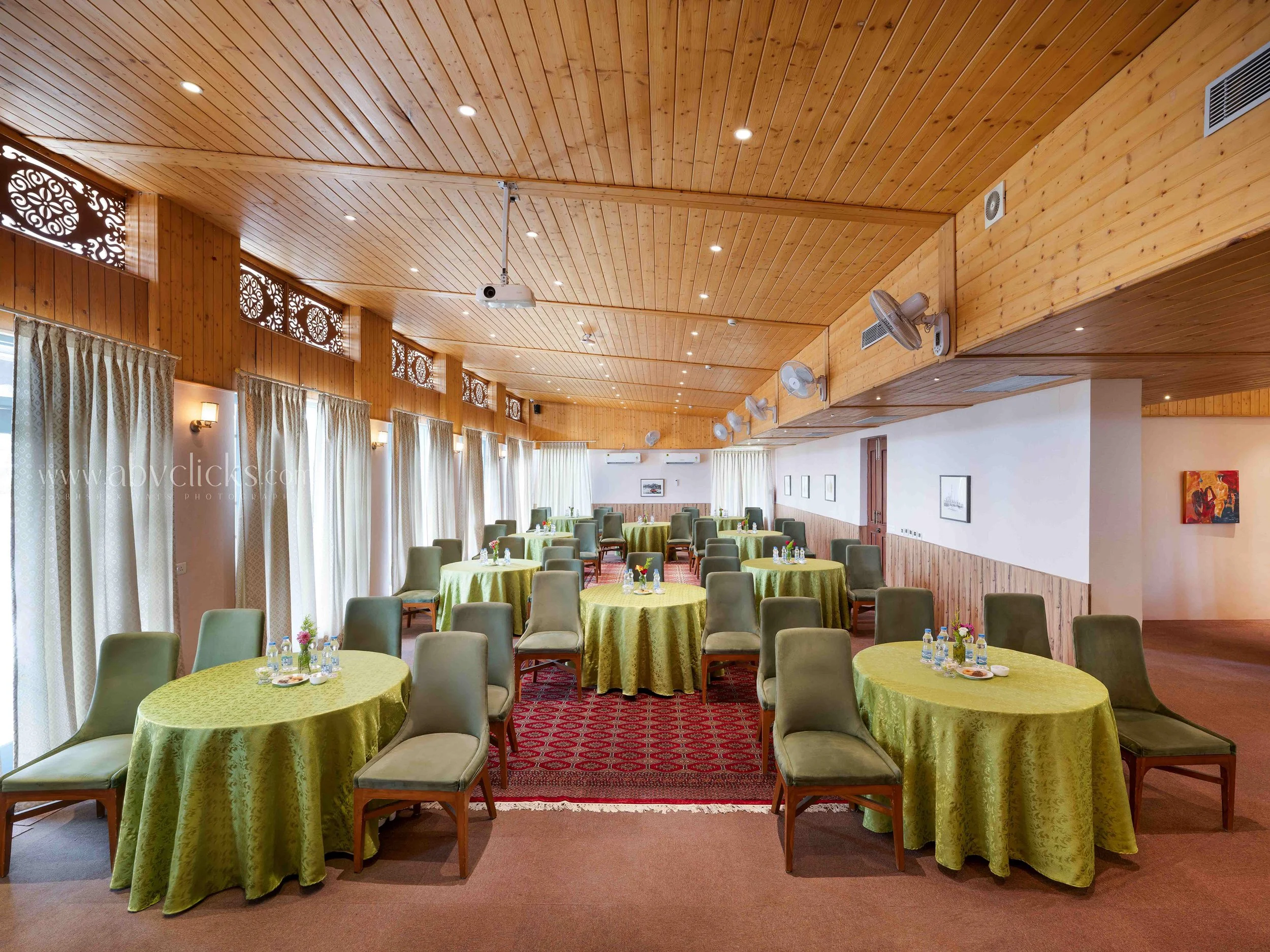 An indoor event room with wooden ceiling and walls, decorated with white curtains and patterned wood panels near windows. Multiple round tables with green tablecloths are set with bottles of water, glasses, and small flower arrangements. The room has