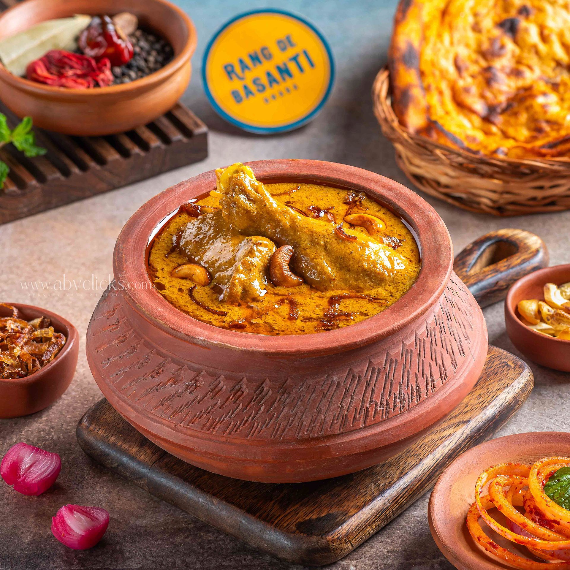 Close-up of an Indian curry with chicken in a clay pot, decorated with cashew nuts, with side bowls of pickles and onion slices, on a rustic wooden board, with a spicy yellow curry labeled 'RAME DE BASANTI' in the background.