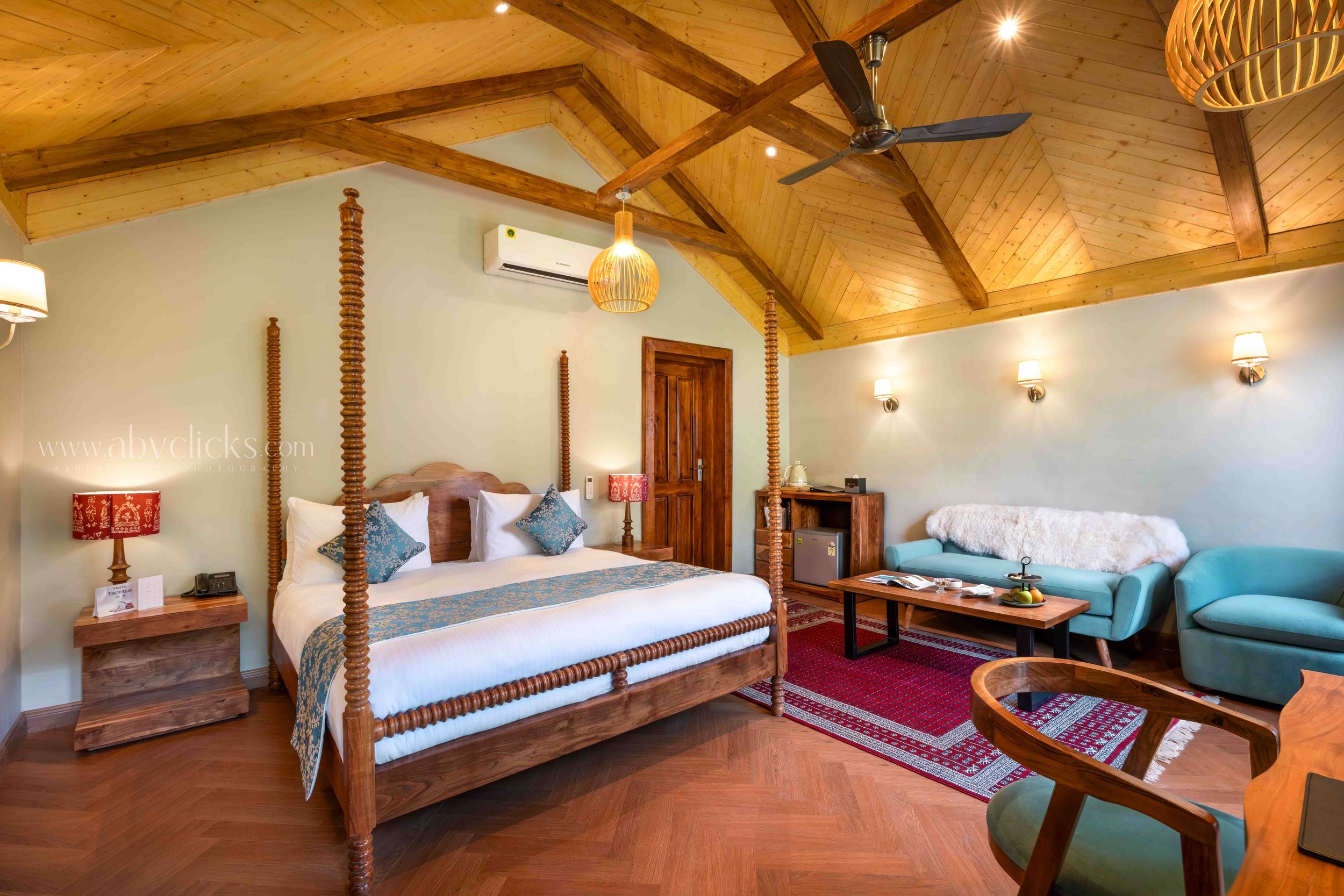 Hotel room with wooden four-poster bed, teal sofa, wooden ceiling, and warm lighting.