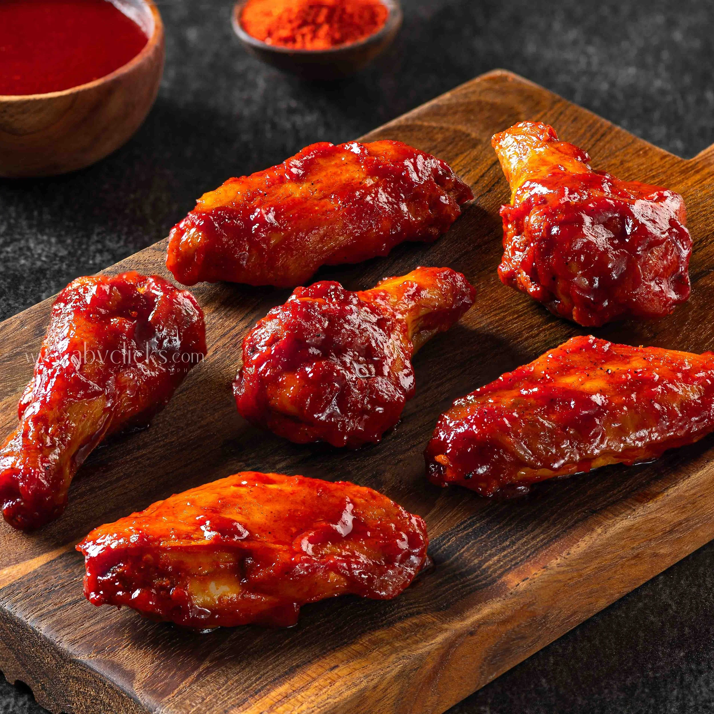 Close-up of glazed chicken pieces with a red sauce on a wooden cutting board, with bowls of red spices or sauce in the background.