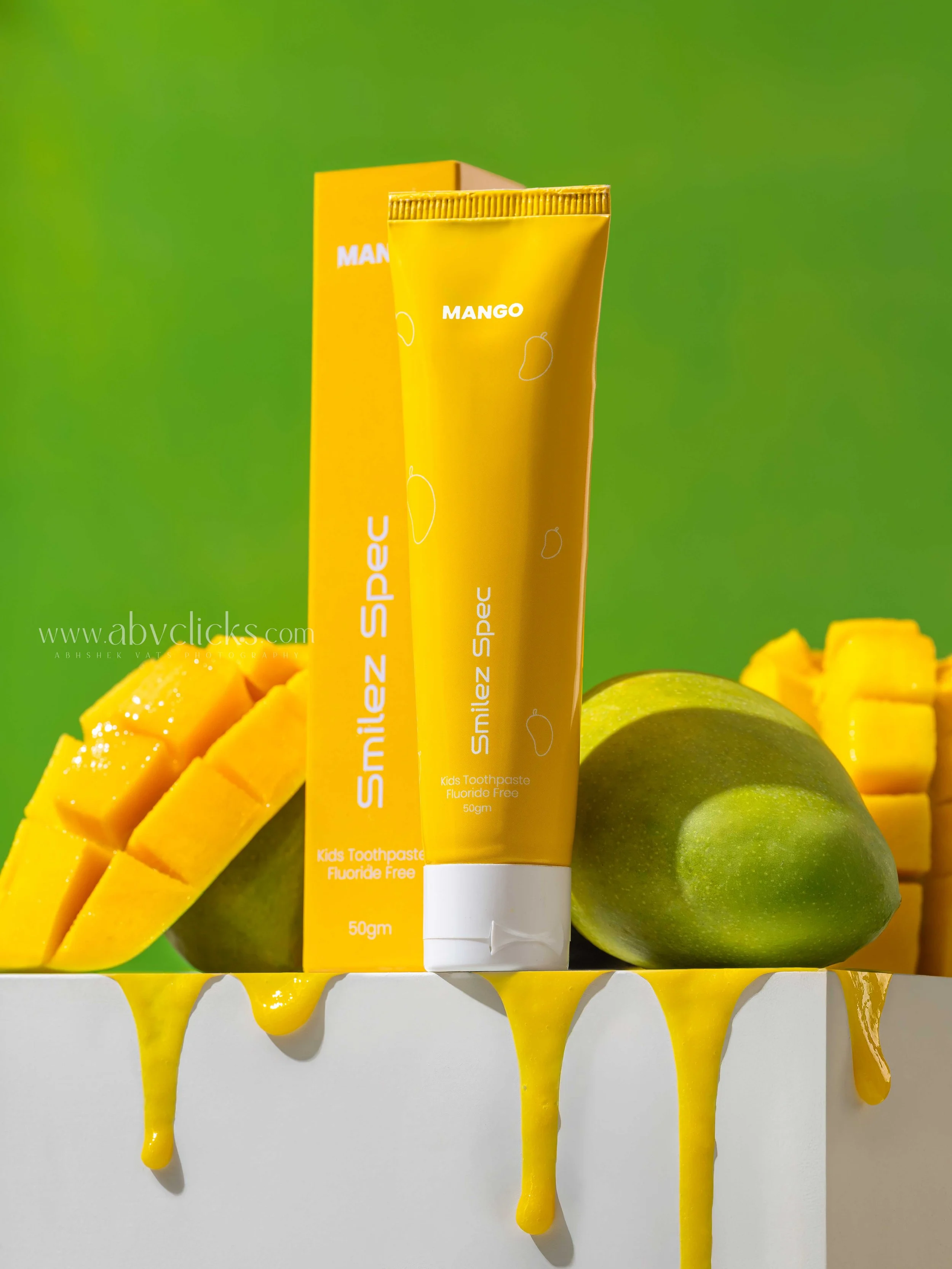 A tube of yellow kids' mango-flavored fluoride-free toothpaste standing upright with its box behind it. The tube and box feature white text and small mango illustrations. The toothpaste is surrounded by cut mango slices and whole green mangoes, with 