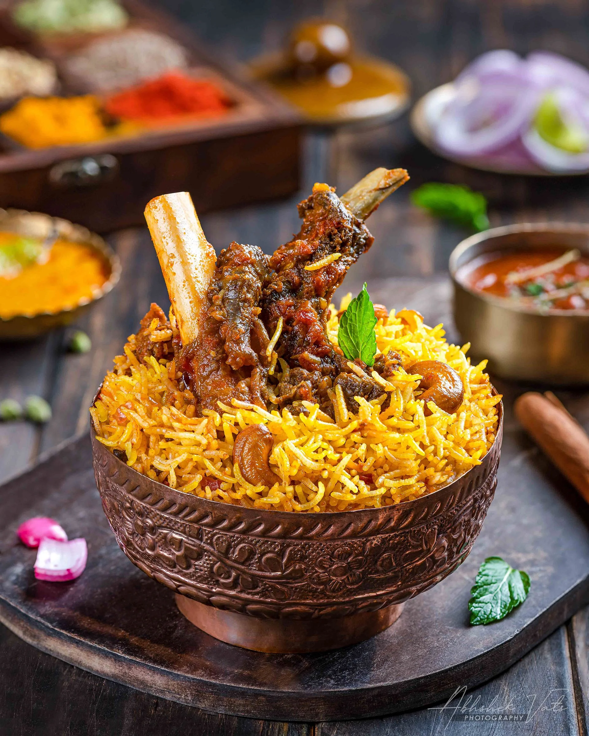 A decorative bowl filled with saffron rice, topped with a cooked lamb chop, garnished with mint leaves, set on a dark wooden tray with scattered flower petals and greenery.