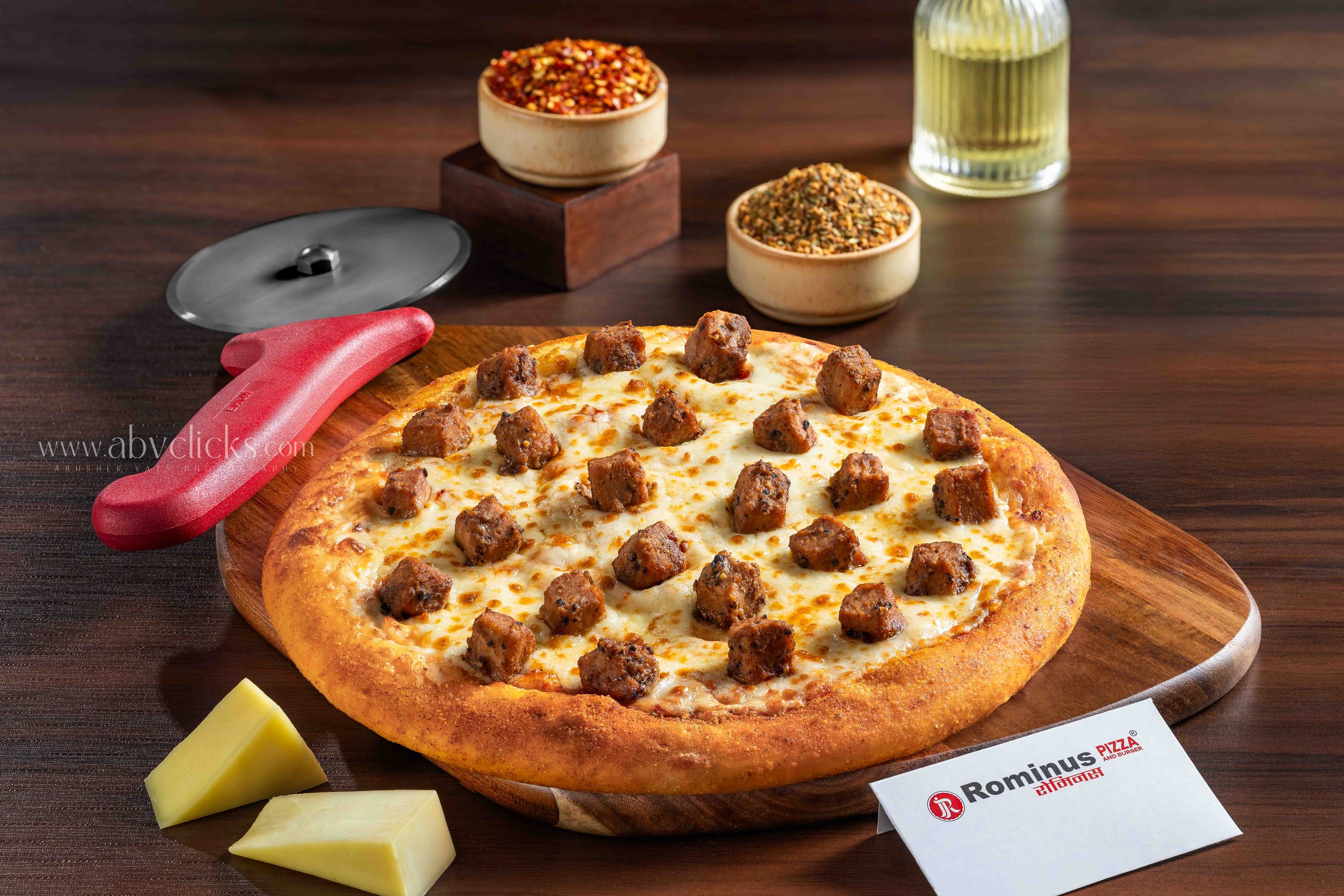 A pepperoni and sausage pizza on a wooden pizza board, with slices of cheese nearby, and bowls of crushed red pepper and other herbs in the background.