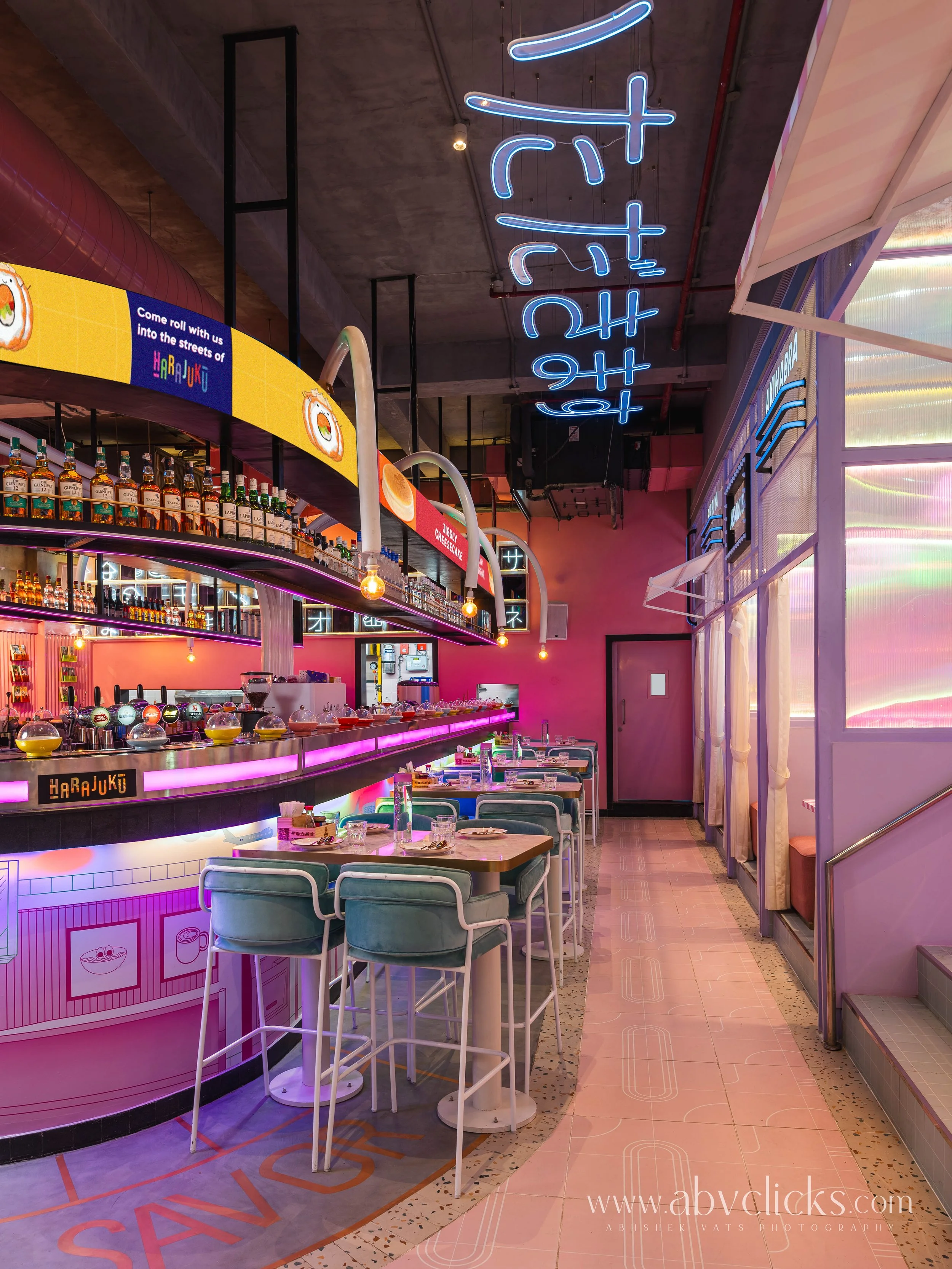Interior of a colorful restaurant with neon signs, pink walls, and modern decor. There are tables and chairs, a bar area with bottles, and a sign above that says 'Harajuku'.