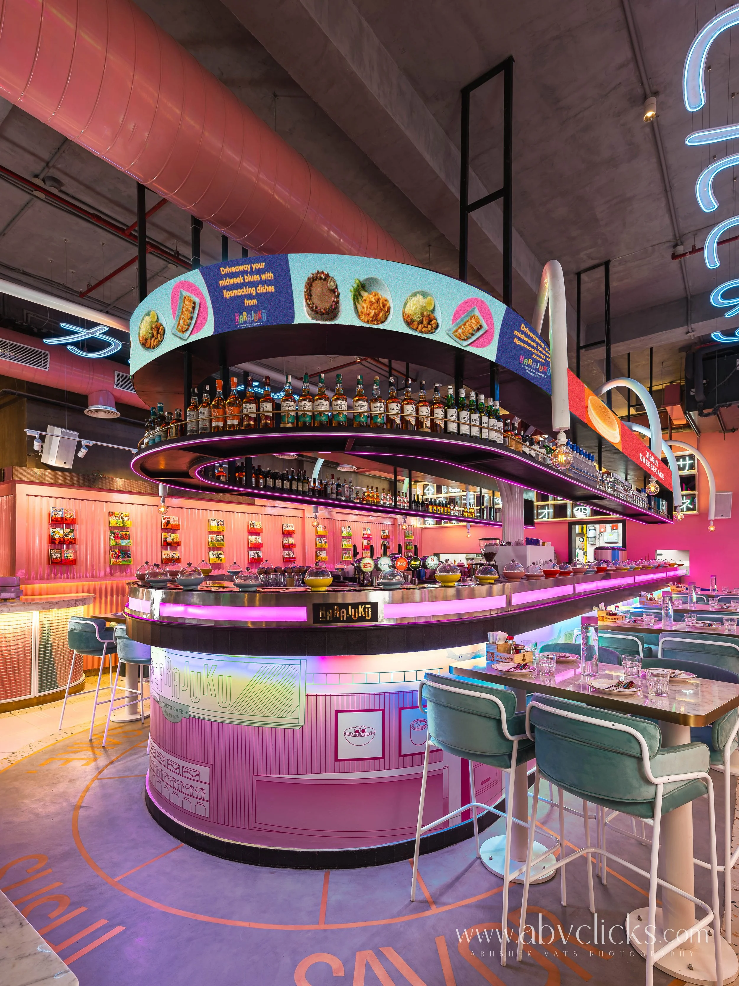 A modern restaurant or cafe with a pink neon illuminated bar, colorful food images displayed on a curved digital signboard, and seating area with pastel-colored chairs and tables.