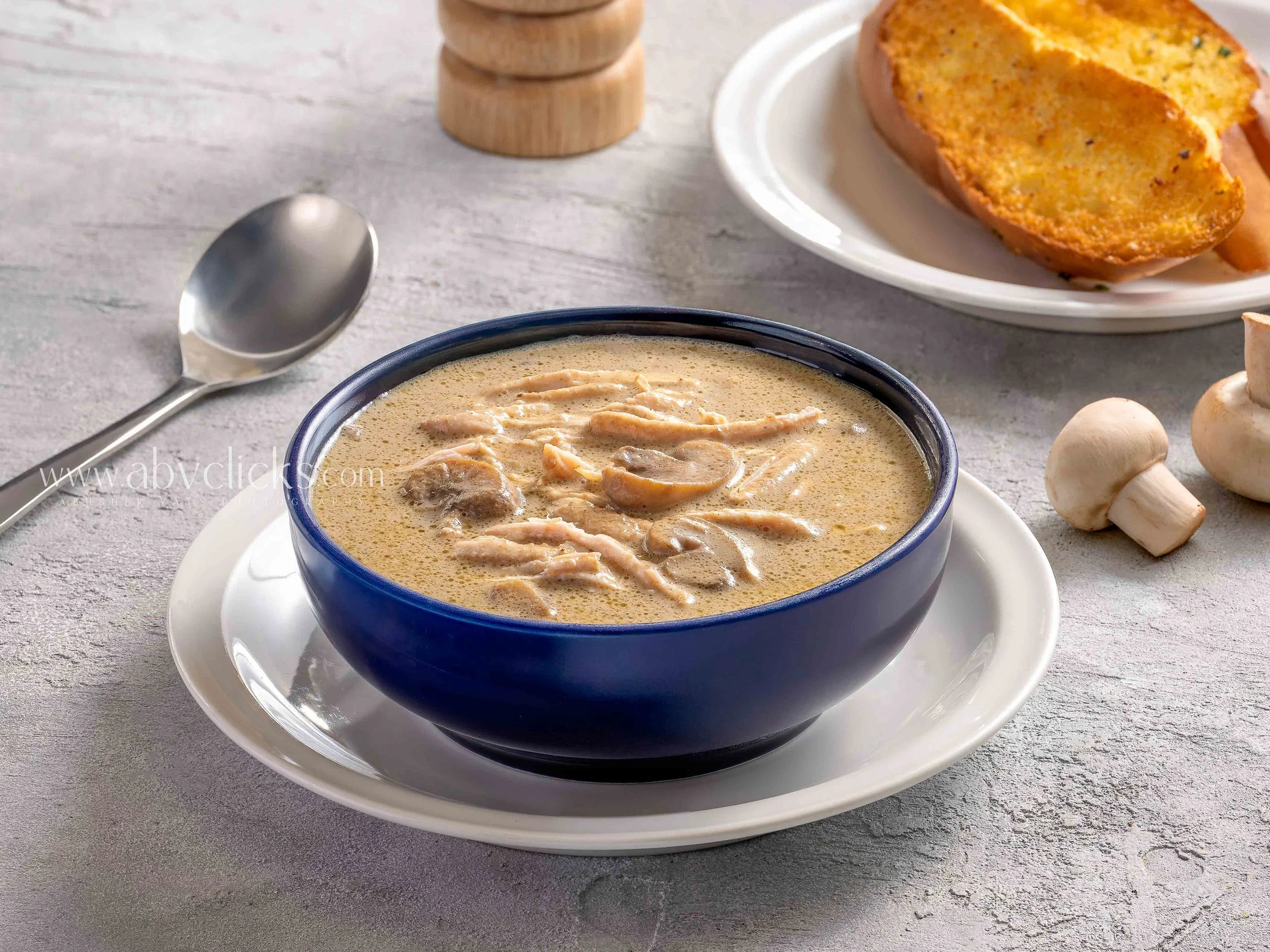 A bowl of creamy chicken and mushroom soup garnished with shredded chicken, served on a white plate with a spoon nearby. In the background, there is a plate with half a toasted garlic bread and some whole mushrooms on a gray wooden surface.