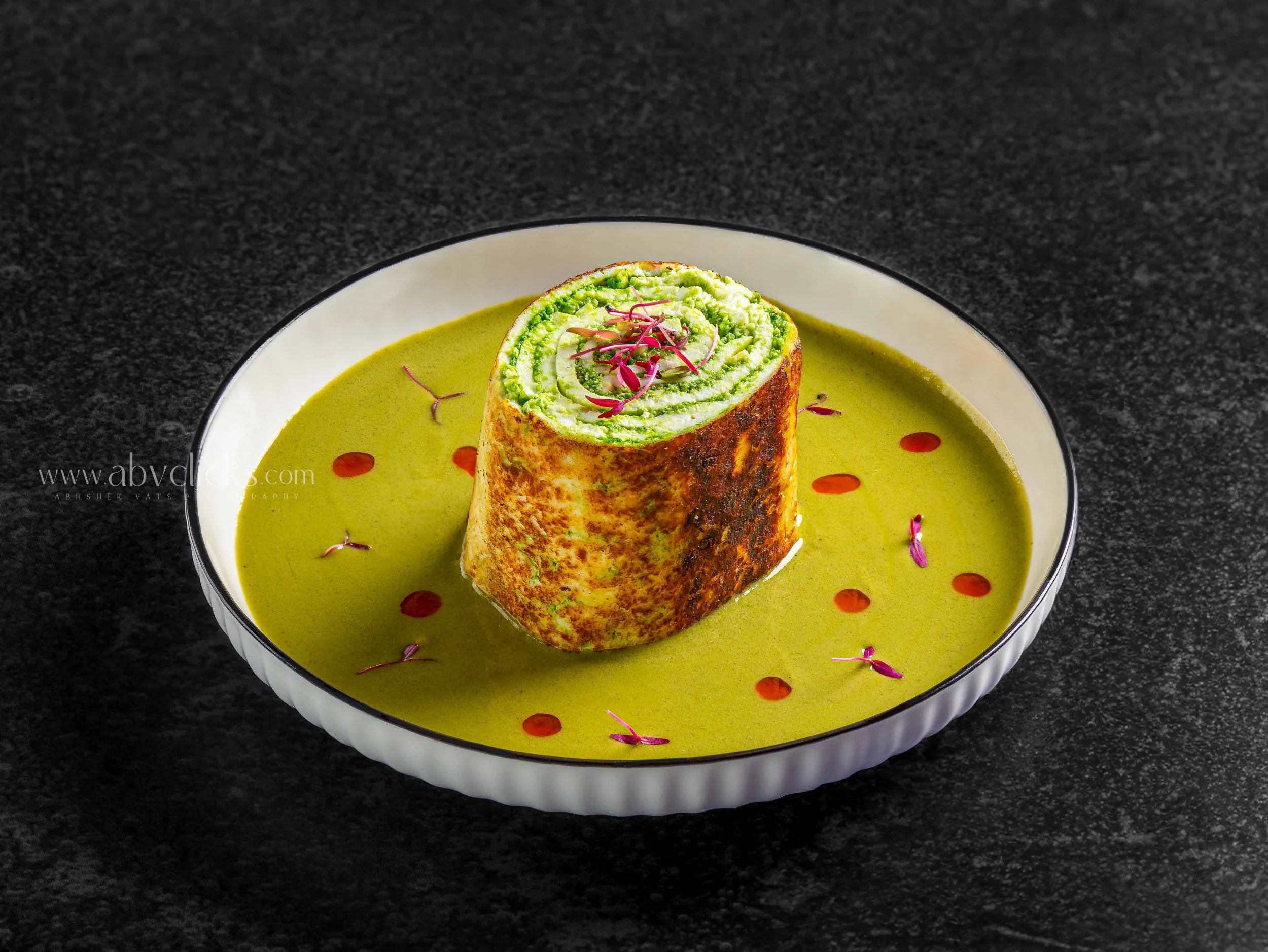 A white bowl with black trim containing green creamy soup garnished with microgreens and red dot accents, with a rolled green filling topped with microgreens on top, set against a dark textured background.