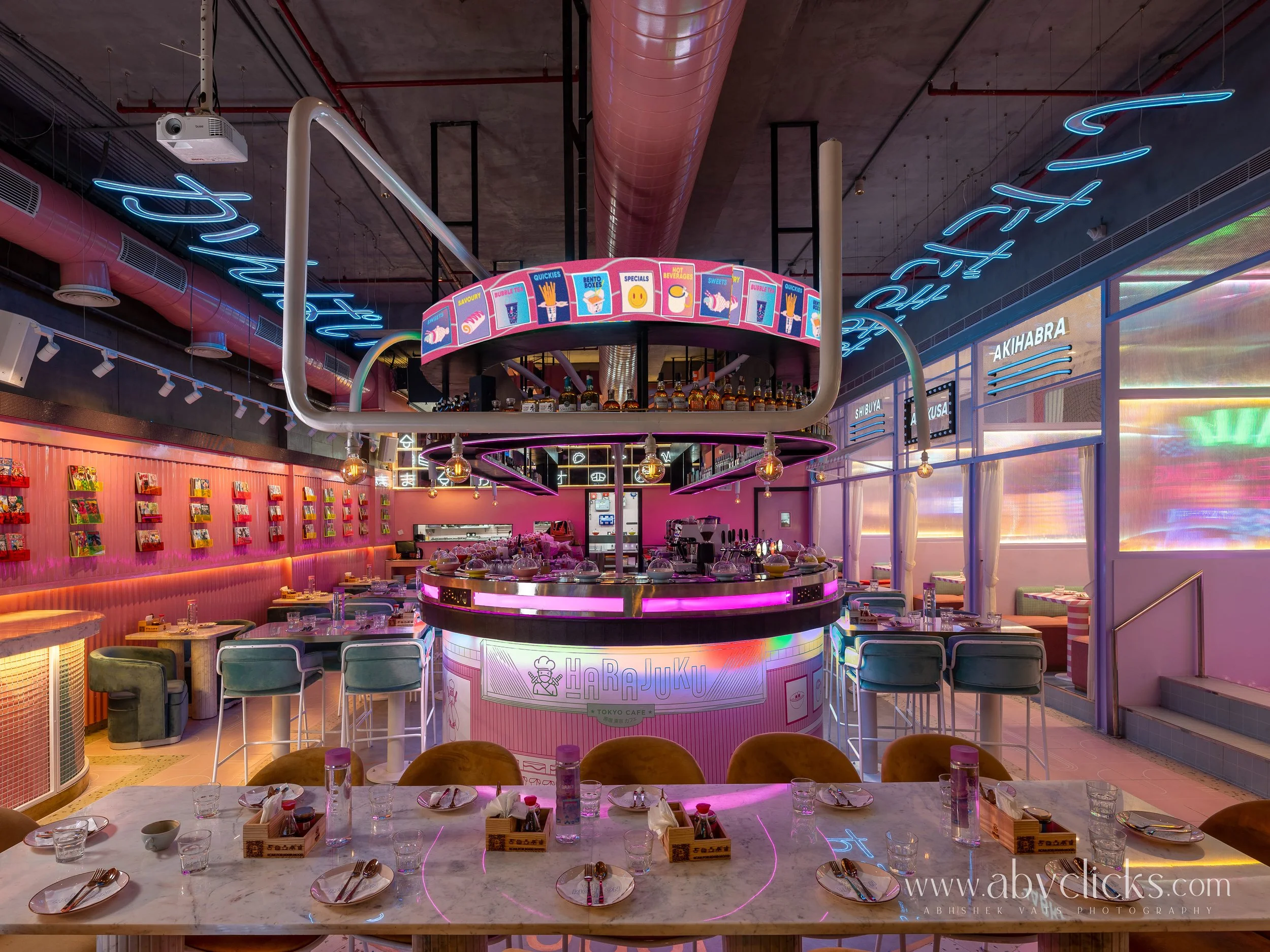 Interior of a modern cafe with pink walls, neon signs, a central pink bar with illuminated signs and bottles, and tables set with plates, glasses, and utensils.