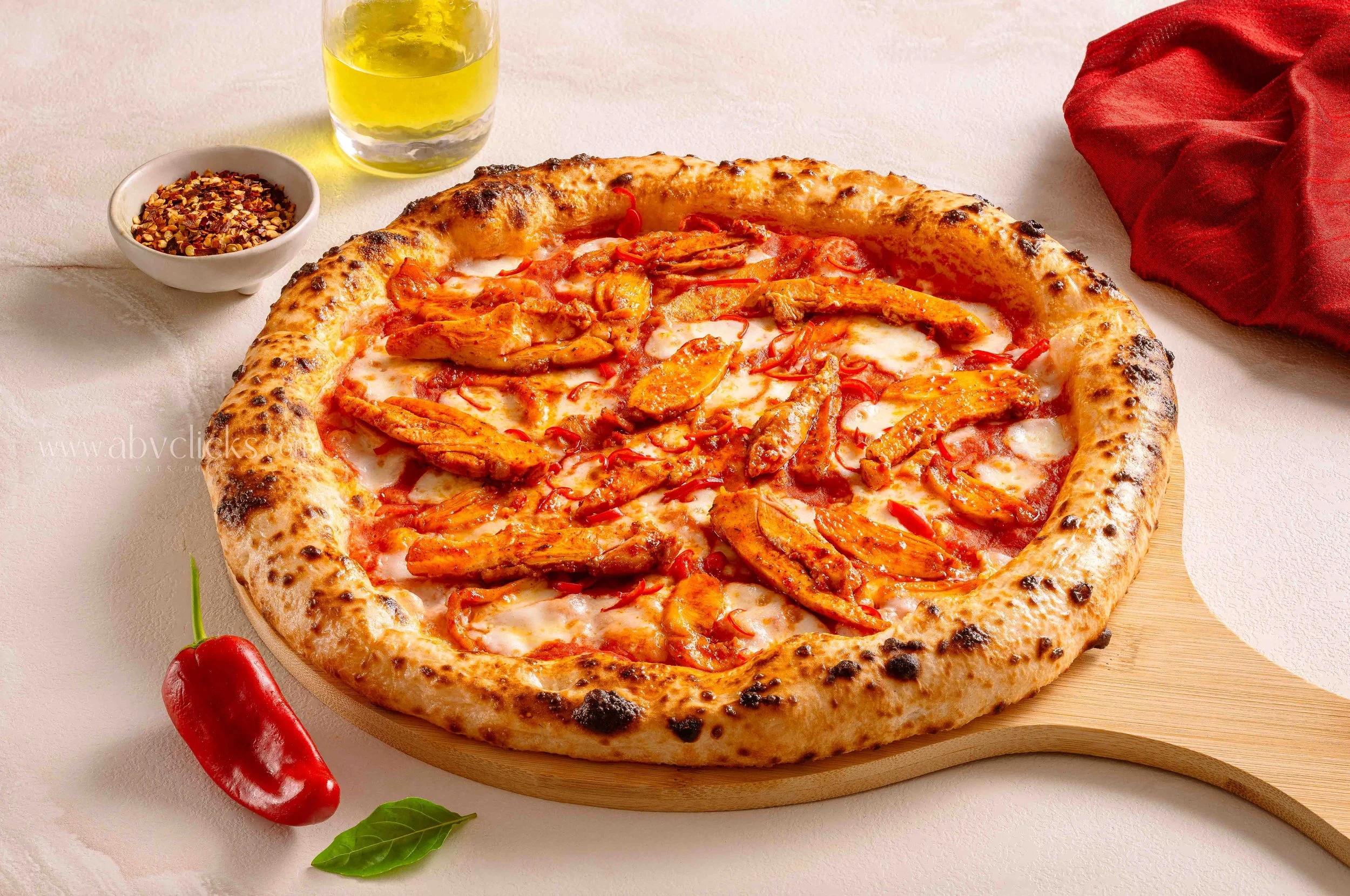 A cooked pizza with chicken, red peppers, and cheese on a round wooden board. A bowl of crushed red pepper flakes, a glass of yellow drinking beverage, a red chili pepper, and basil leaves surrounding the pizza.