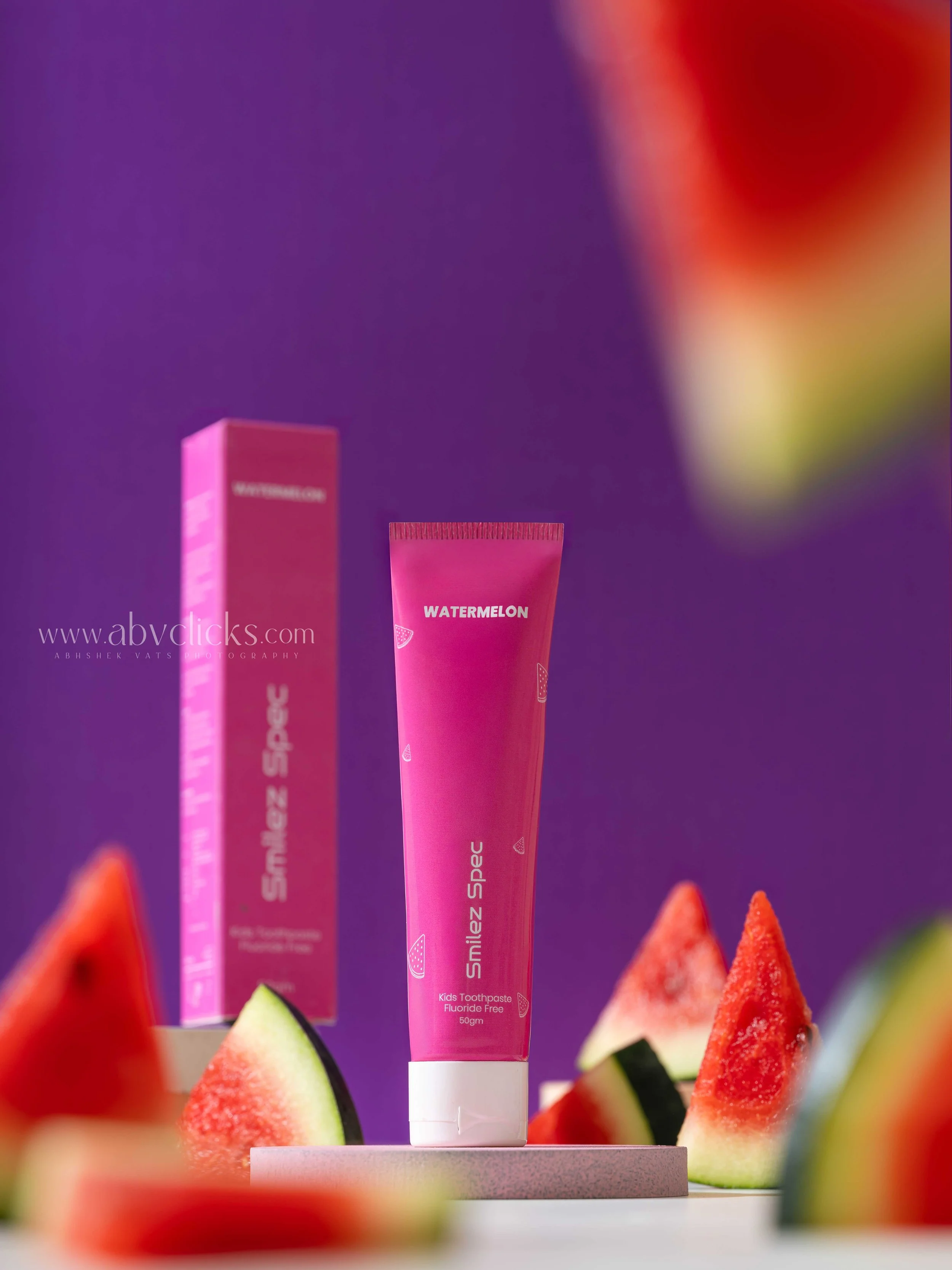 Pink Kids toothpaste tube labeled "Watermelon" and its box, with watermelon slices around it, set against a purple background with blurred red and yellow shapes.