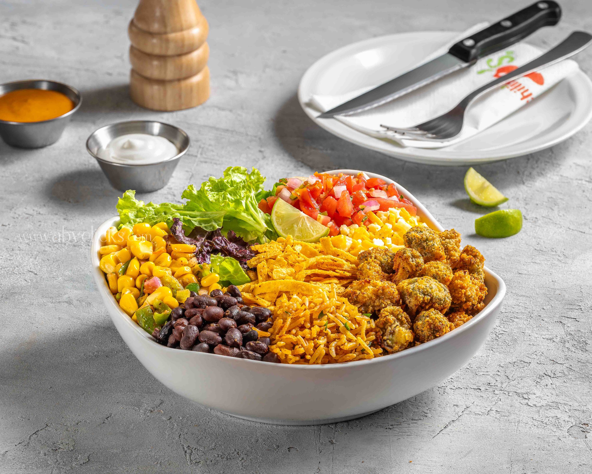 A large white bowl filled with a salad containing lettuce, corn, black beans, chopped tomatoes, shredded cheese, and fried food balls, with lime wedges on the side. In the background, there are small metal bowls with orange sauce and sour cream, a wo